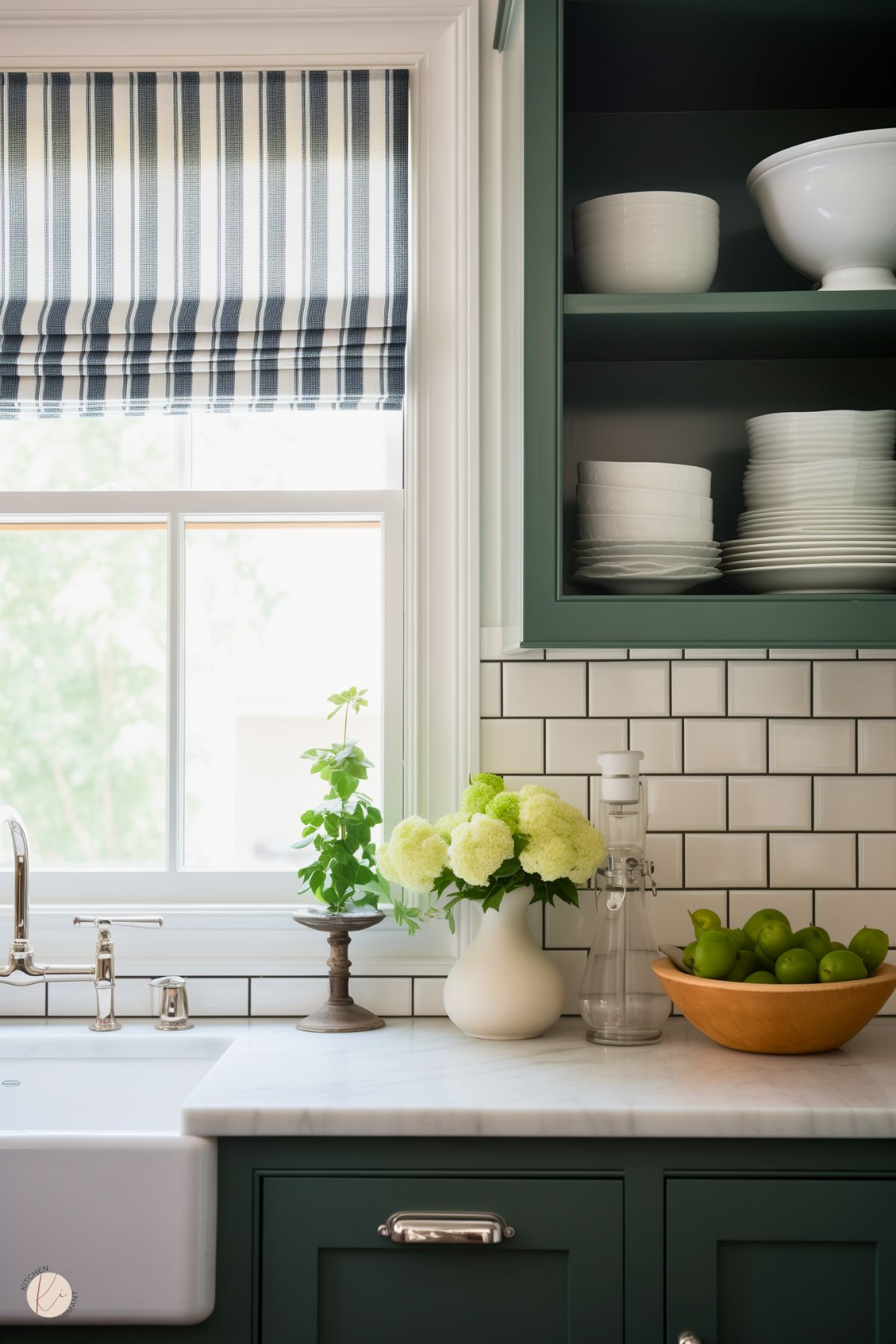 A cozy preppy kitchen corner with deep green cabinetry, a white subway tile backsplash, and marble countertops. The striped Roman shade, fresh green apples, and floral arrangement add charm and natural elegance to the space.