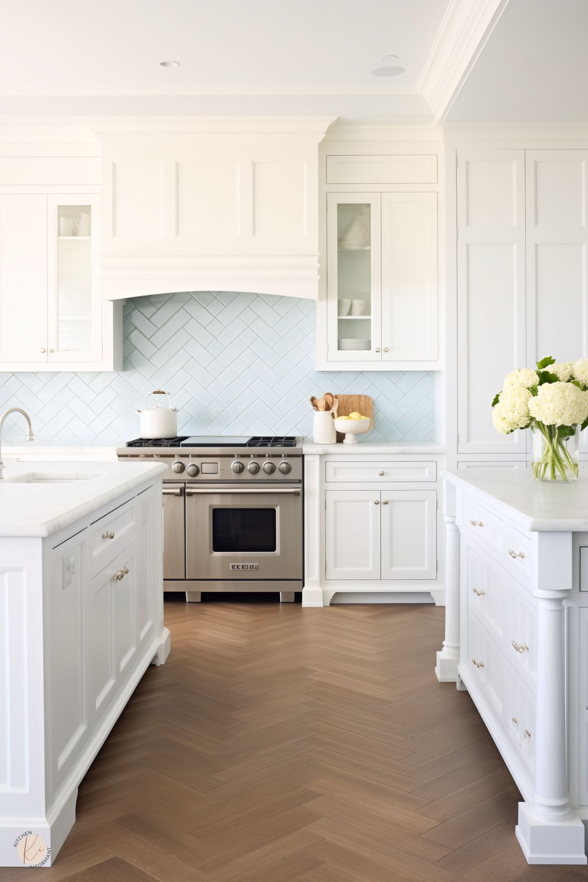 A crisp preppy kitchen with white cabinetry, a light blue herringbone tile backsplash, stainless steel appliances, and marble countertops. The warm wood herringbone flooring adds a touch of elegance, complemented by fresh floral accents.