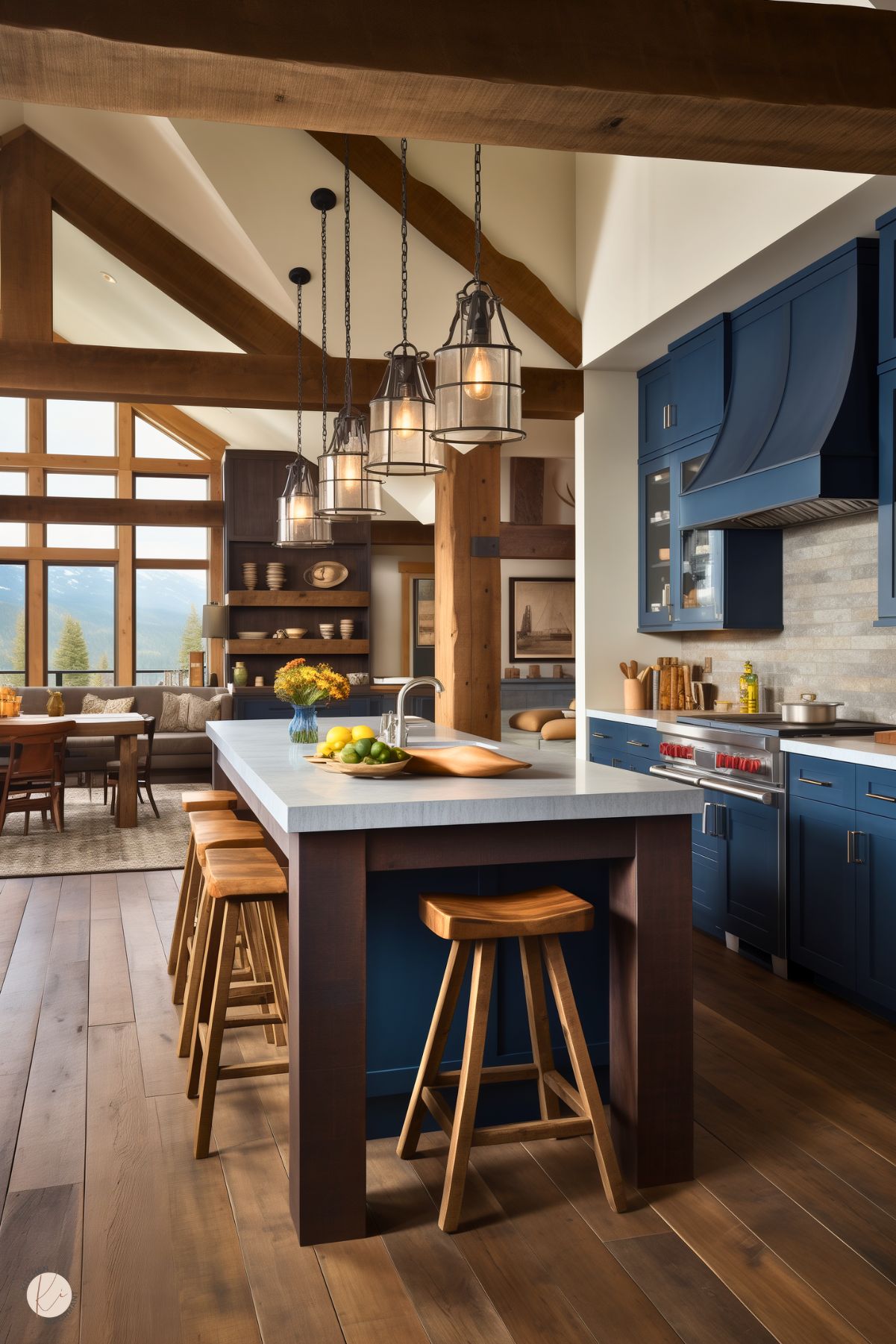 A spacious rustic kitchen with navy blue cabinetry, a large island with a white countertop, and warm wood accents. The island features dark wood legs and matching wooden barstools, with a bowl of fresh fruit adding a natural touch. Lantern-style pendant lights with metal framing hang above. The open-concept space extends into a cozy dining and living area, with large windows offering stunning mountain views. Exposed wooden beams and a textured backsplash enhance the rustic charm.