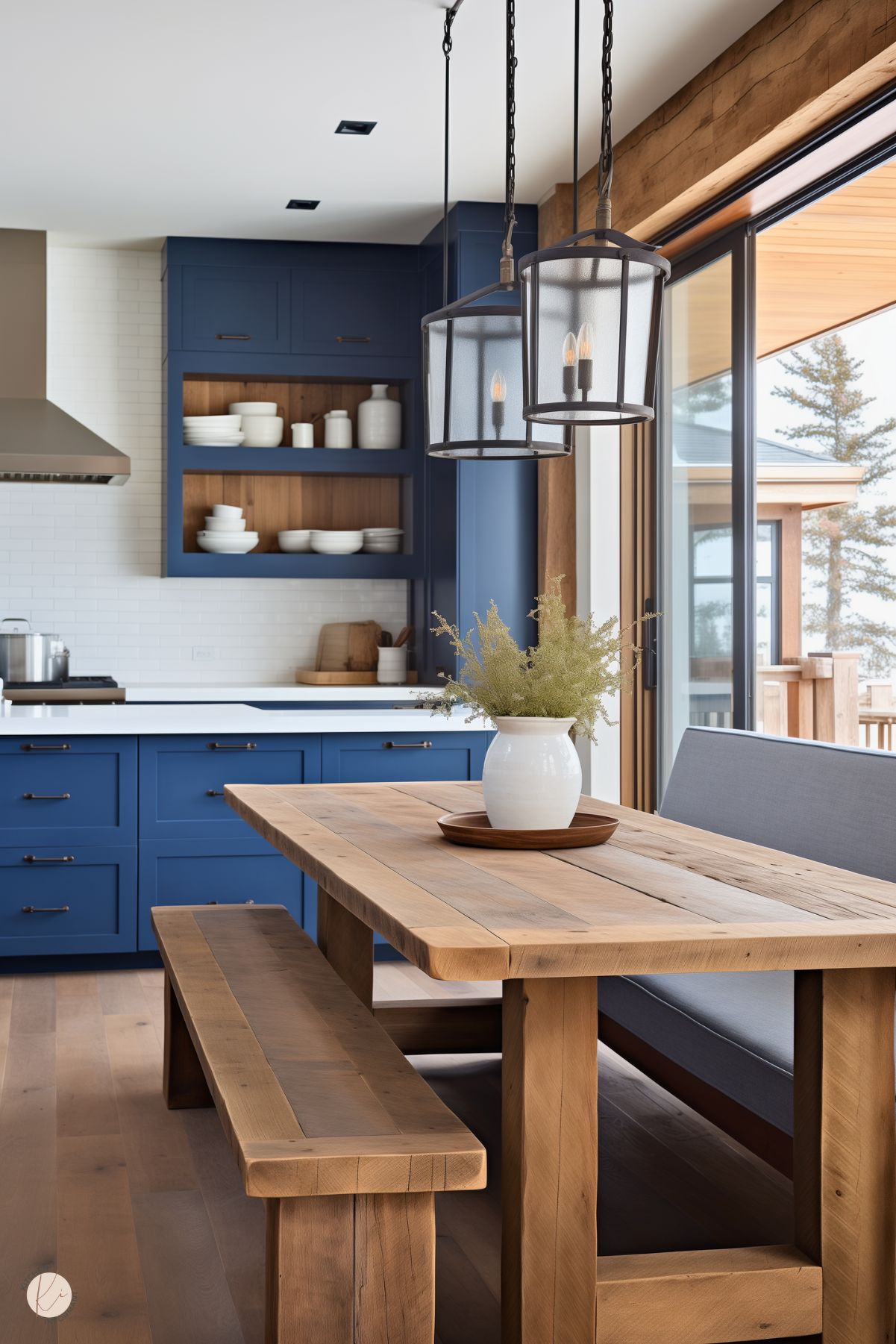 A rustic kitchen and dining area with navy blue cabinetry, black hardware, and a white subway tile backsplash. Open wooden shelving with a blue frame holds neatly arranged white dishware. A large wooden farmhouse table with a matching bench and upholstered seating sits under two black metal lantern-style pendant lights. A white vase with greenery serves as a simple centerpiece. Large sliding glass doors lead to an outdoor deck, letting in natural light and offering a scenic view.