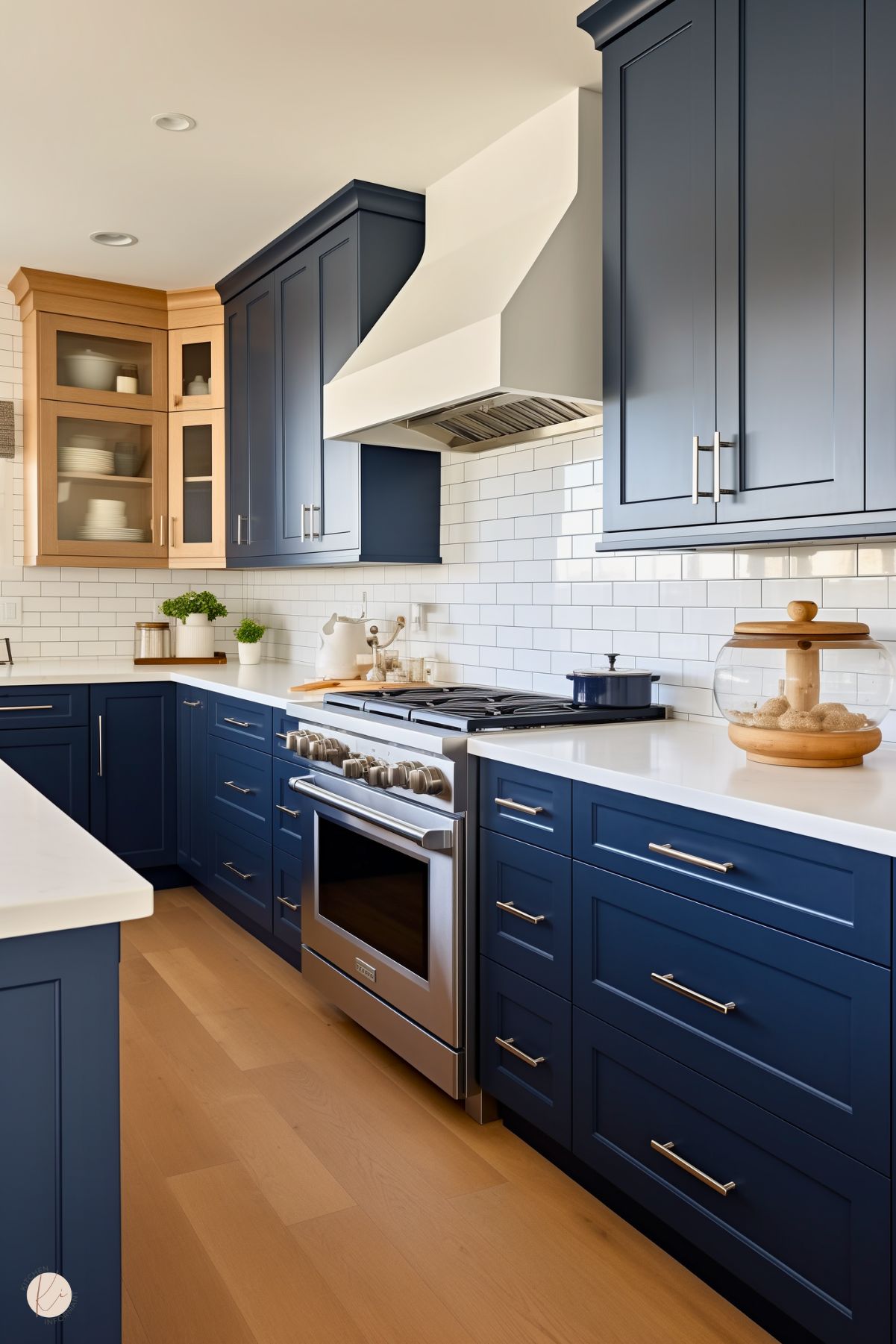 A kitchen with navy blue cabinets, white quartz countertops, and a white subway tile backsplash. A mix of wood and glass-front cabinetry adds warmth, while a custom white range hood blends seamlessly with the design. Stainless steel appliances and brushed nickel hardware complete the modern rustic look. Light wood flooring enhances the bright and inviting space.