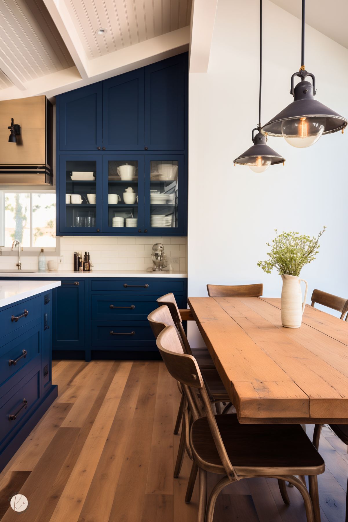 A rustic kitchen with navy blue cabinetry, glass-front upper cabinets, and a white subway tile backsplash. A warm wood dining table with matching chairs sits nearby, accented by industrial pendant lights. The space features a vaulted ceiling with exposed beams, a brass range hood, and natural light from a nearby window, creating a cozy yet refined look.