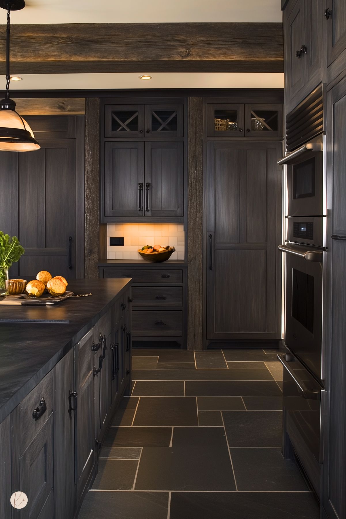 A modern kitchen with deep-toned cabinetry and slate tile flooring. The wood grain of the dark cabinets is complemented by black metal hardware. A built-in double oven and illuminated backsplash add functionality and warmth. The central island features a matching dark surface and holds a bowl of fruit and bread for a welcoming touch. Exposed wooden ceiling beams enhance the space’s cozy, rustic feel.