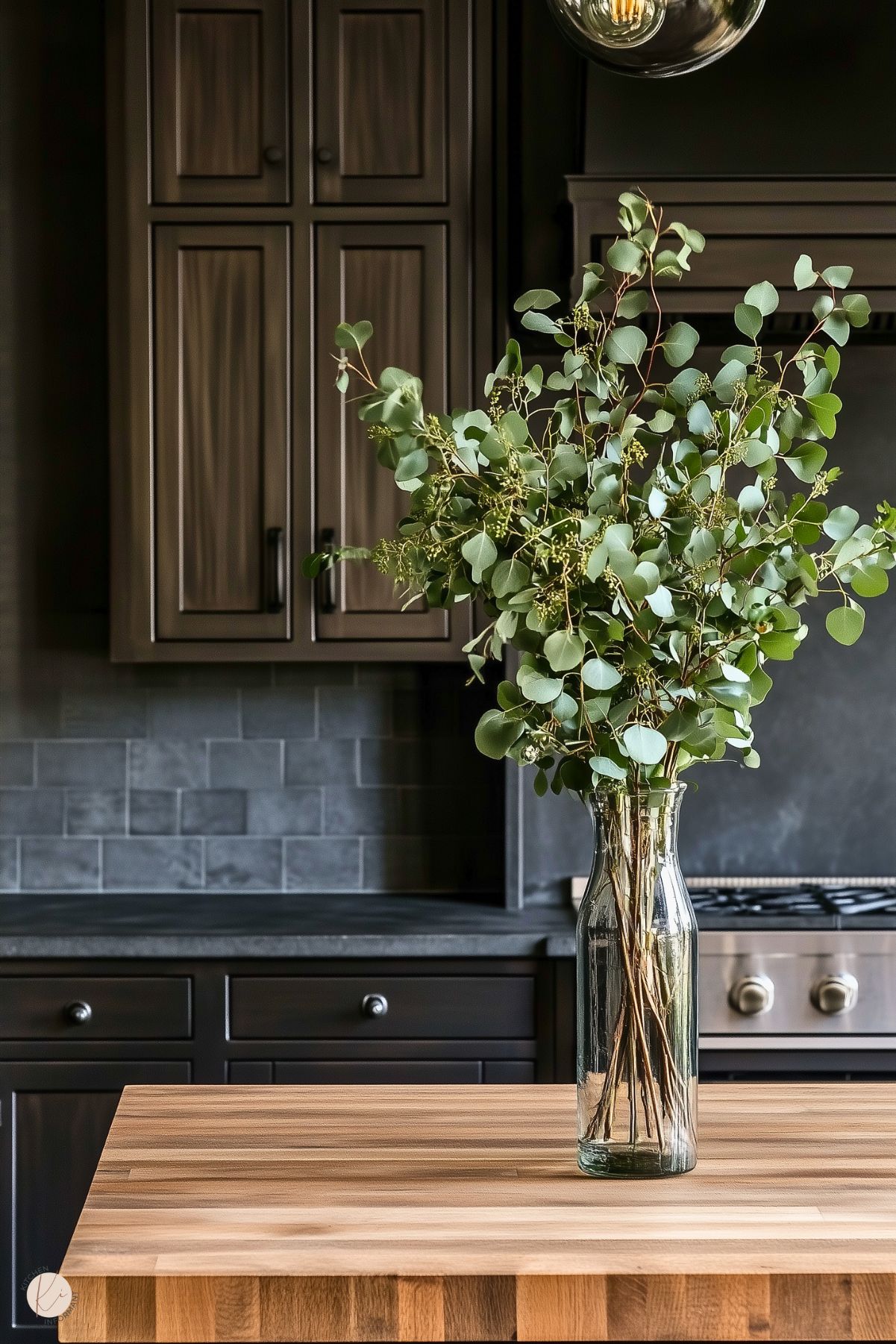 A close-up view of a kitchen island featuring a butcher block countertop with a clear glass vase holding a tall arrangement of eucalyptus branches. The background showcases dark cabinetry, a subtle tiled backsplash in gray tones, and part of a stainless steel stovetop, creating a cozy yet elegant atmosphere with earthy and modern elements.