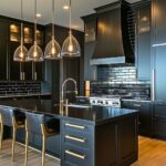 A sophisticated kitchen featuring matte black cabinetry, a matching black island, and a striking black range hood. Gold hardware and fixtures, including sleek handles and a modern faucet, add a touch of elegance. The island is illuminated by four glass pendant lights with warm bulbs, creating a cozy ambiance. The backsplash features glossy black subway tiles, adding depth and texture. The light wood flooring provides a warm contrast to the dark tones, tying the space together beautifully.