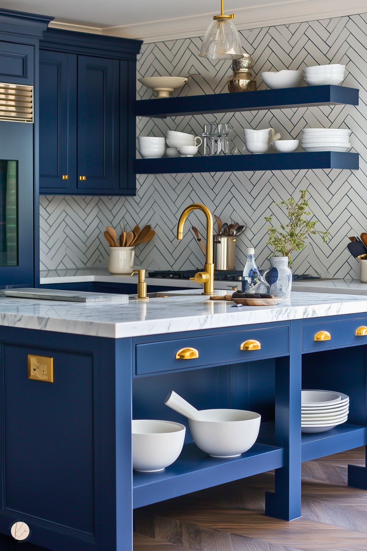 A bright kitchen featuring navy blue cabinetry and open shelving, accented with gold hardware and fixtures. The marble countertop and herringbone backsplash in white add a classic touch, while the gold faucet stands out as a luxurious detail. The island includes open shelving for white dishware, and a decorative vase with greenery adds a fresh, natural element. Warm wood flooring grounds the modern, elegant design.