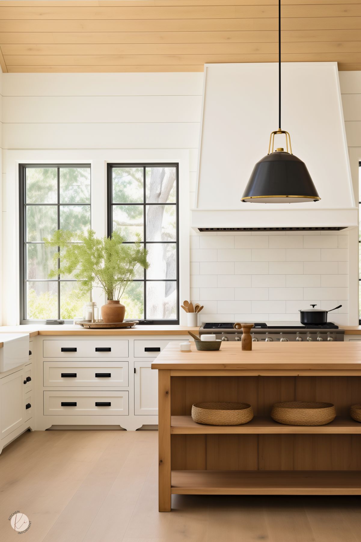 A cozy farmhouse kitchen with white cabinetry, black hardware, and a warm wood island featuring open shelving with woven baskets. The backsplash is finished in classic white subway tile, complemented by a large white range hood. Natural light pours in through black-framed windows, highlighting a terracotta vase with greenery. A black pendant light with brass accents hangs above the island, completing the inviting look.