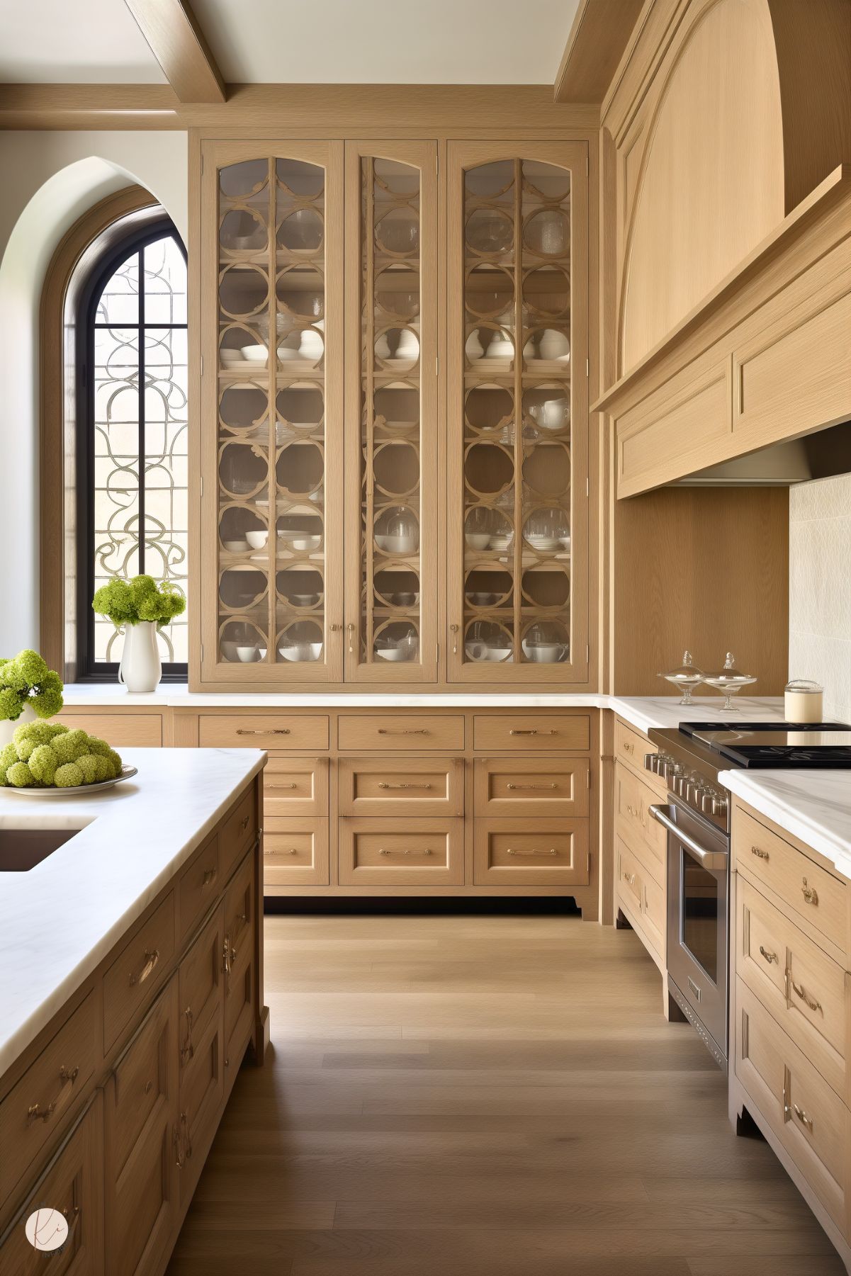 A classic kitchen featuring white oak cabinetry with intricate glass-paneled doors displaying white dishware. The island and countertops are topped with polished white stone, complemented by brass hardware. An arched window with ornate detailing brings in natural light, while light wood floors and a built-in range complete the sophisticated design. A vase of green hydrangeas adds a pop of color.