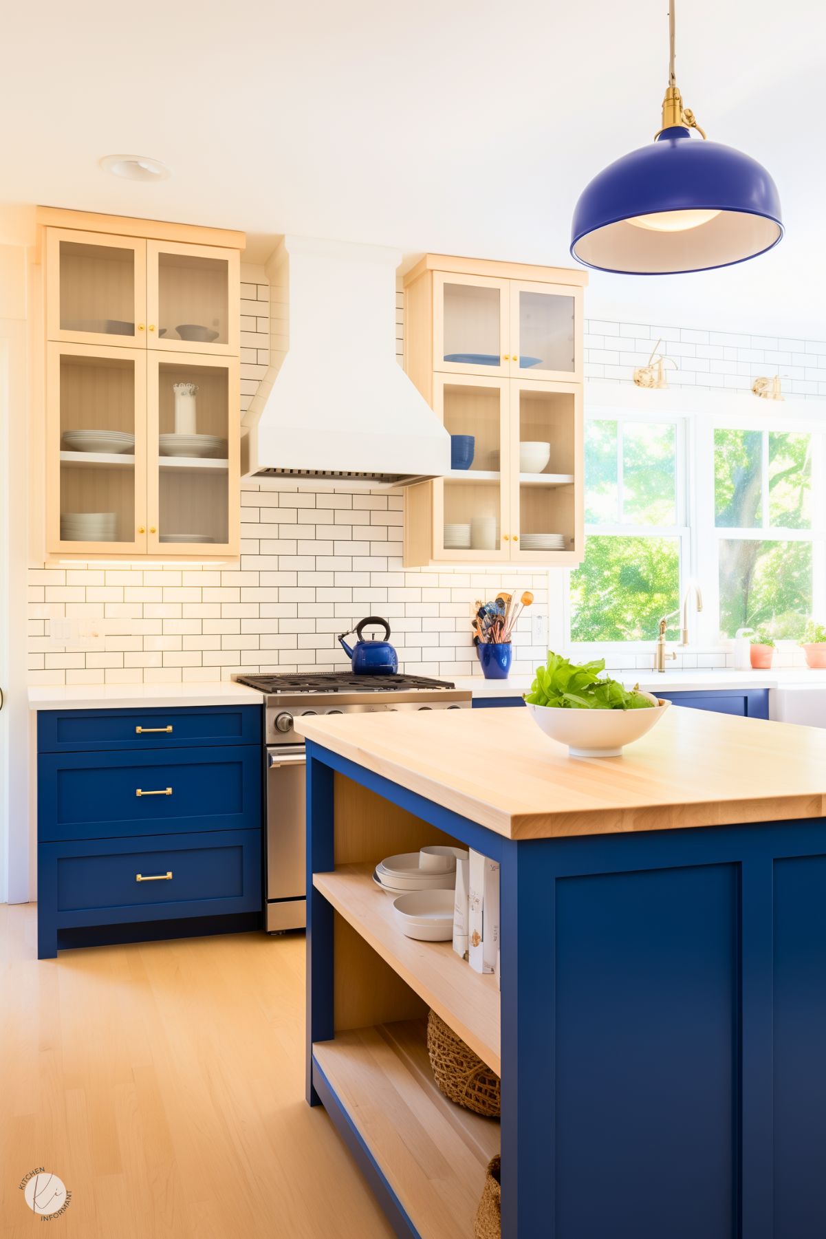 A bright and inviting kitchen with bold blue lower cabinets and a matching island featuring a light wood butcher block countertop. Upper cabinets with glass fronts and a cream finish complement the white subway tile backsplash. A blue pendant light above the island adds a cohesive accent, while natural light streams in through large windows, highlighting the airy and fresh design. A blue kettle and pops of green from fresh produce complete the look.