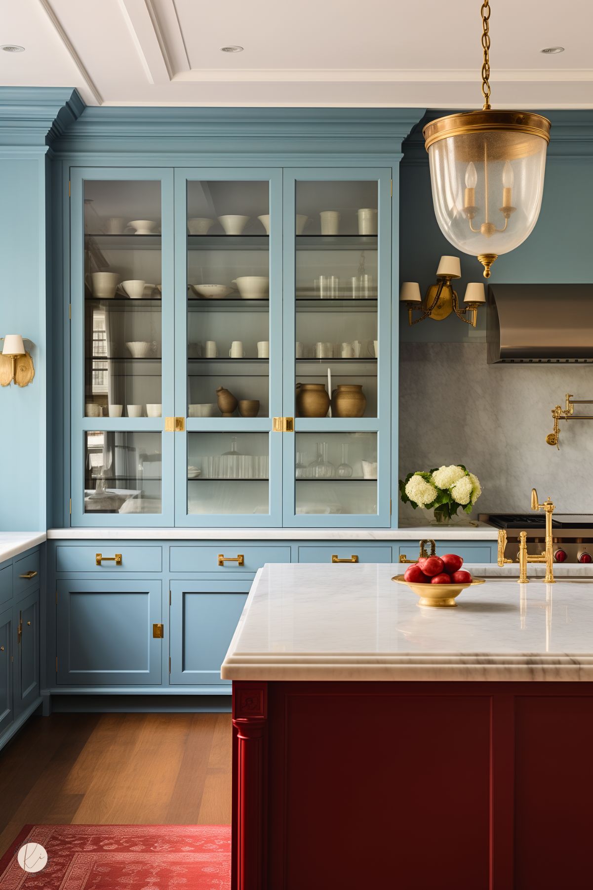 A luxurious kitchen featuring powder blue cabinetry with glass-front doors and gold hardware. The central island stands out in a rich red tone, topped with a white marble countertop adorned with a gold fruit bowl filled with red apples. Gold fixtures, including a faucet and pendant light, add an elegant touch, complemented by a marble backsplash and stainless steel range hood. A red rug and floral arrangement complete the sophisticated, classic design.