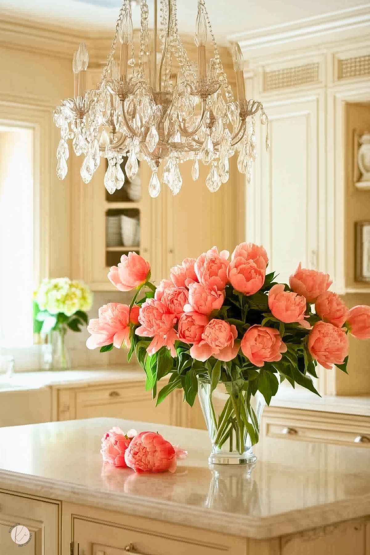 A kitchen with cream-colored cabinetry and a marble countertop island. A crystal chandelier hangs above the island, adding a decorative focal point. The island is adorned with a vase of vibrant pink peonies, and additional greenery is visible in the background. The cabinetry features paneled details, and natural light enters through a nearby window, enhancing the soft, neutral tones.