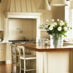 A kitchen with cream-colored cabinetry, a wood countertop island, paneled detailing, and hardwood flooring. The space includes two beige pendant lights above the island, rustic barstools, a vase with white roses, and a range with a built-in pot filler under a decorative hood. Natural light enters through a large window, highlighting the neutral tones.