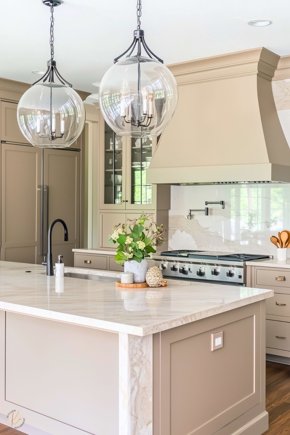 A warm-toned kitchen featuring soft taupe cabinetry and a large marble-topped island with a built-in black faucet. Two oversized glass pendant lights hang above the island. A gas stove with a sleek stainless steel hood is centered against a marble backsplash. A simple floral arrangement and decor pieces add subtle charm.