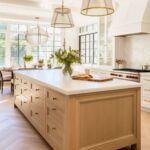 A spacious kitchen with a white oak island featuring a white stone countertop and ample drawer storage with brass hardware. Oversized pendant lights with gold accents hang above, adding warmth to the space. White cabinetry, a herringbone tile backsplash, and large black-framed windows enhance the bright, airy feel. A cozy dining nook with a round table and greenery-filled vases adds charm to the inviting design.