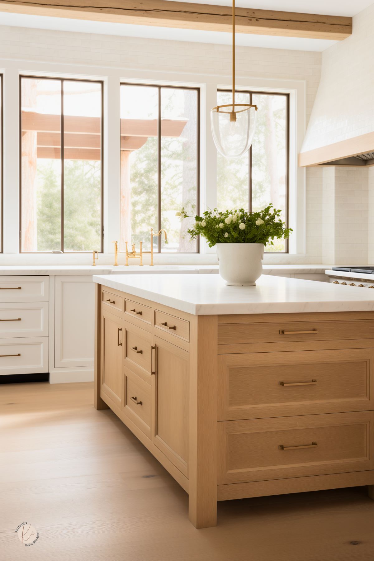 A bright kitchen with a white oak island featuring a white stone countertop and brass hardware. A large glass pendant light with gold accents hangs above, complementing the brass faucet and fixtures. White cabinetry and a subtle tile backsplash create a clean, modern look. Large windows with black metal frames let in natural light, while exposed wood ceiling beams add warmth. A potted plant brings a fresh, organic touch.