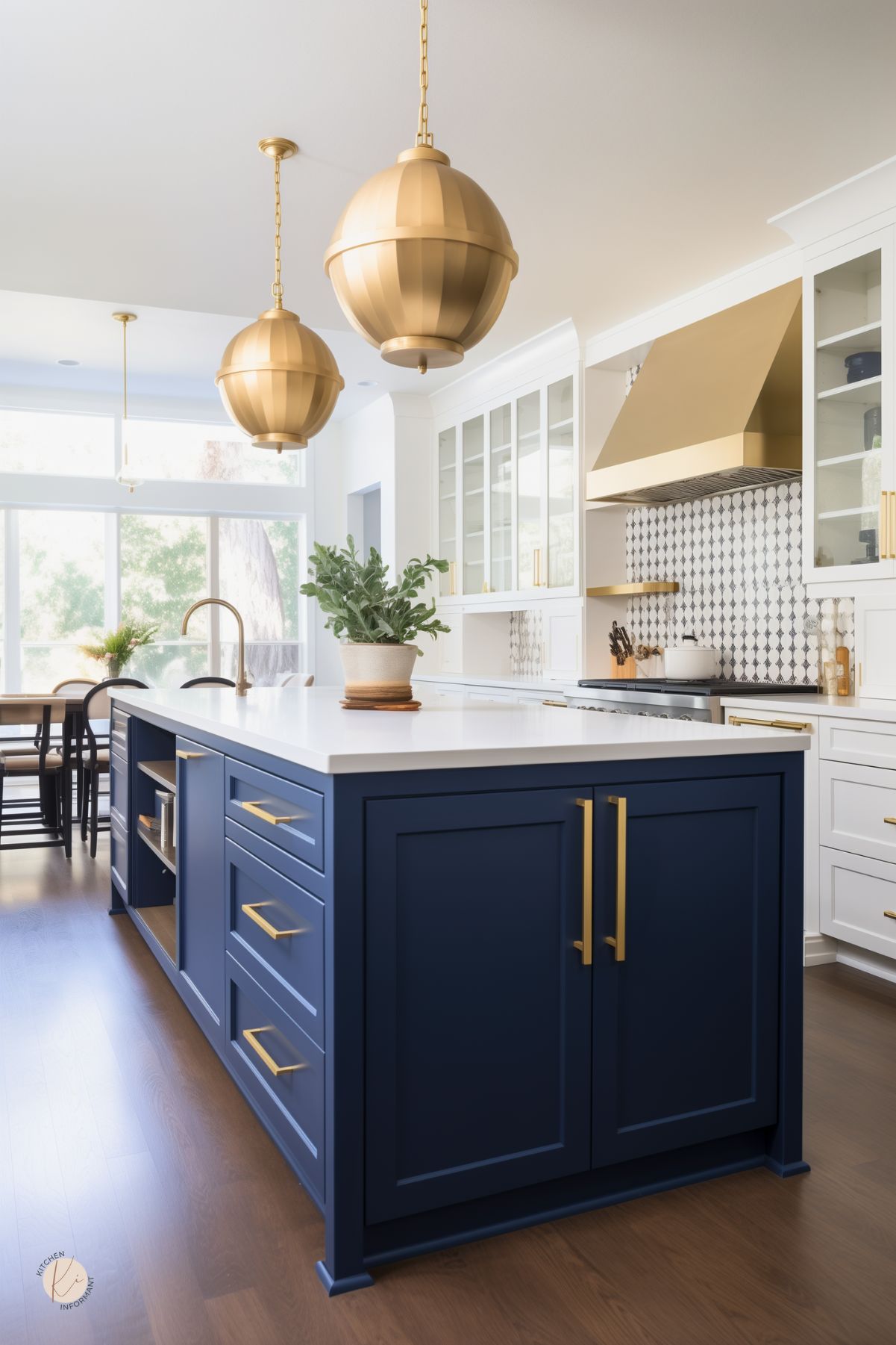 A kitchen with a deep blue island, gold hardware, and a white countertop. Large gold pendant lights hang above, complementing a gold range hood and geometric tile backsplash. White cabinetry with glass doors adds an airy feel, while dark wood flooring contrasts with the bright space. A dining area with black chairs and a large window brings in natural light.