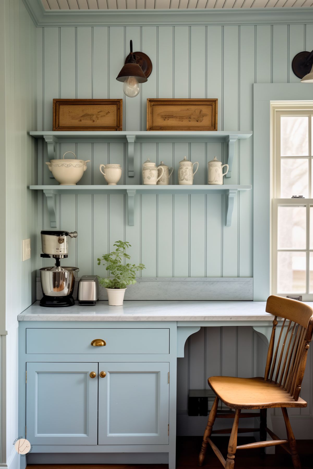 A kitchen nook with soft blue beadboard paneling and a built-in workstation has a cozy, vintage feel. Open shelves hold delicate white dishware and framed antique prints. A marble countertop extends into a small desk area with a wooden spindle-back chair. A stand mixer and toaster sit neatly on the counter, while a potted herb adds a fresh touch. Industrial-style wall sconces provide warm lighting against the pastel backdrop.