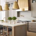 A taupe kitchen with bold brass accents, featuring a statement brass range hood and matching pendant lights. The island has a marble countertop, brass hardware, and wood details, complemented by black upholstered barstools. A sleek marble backsplash and gold pot filler complete the elegant yet modern design.