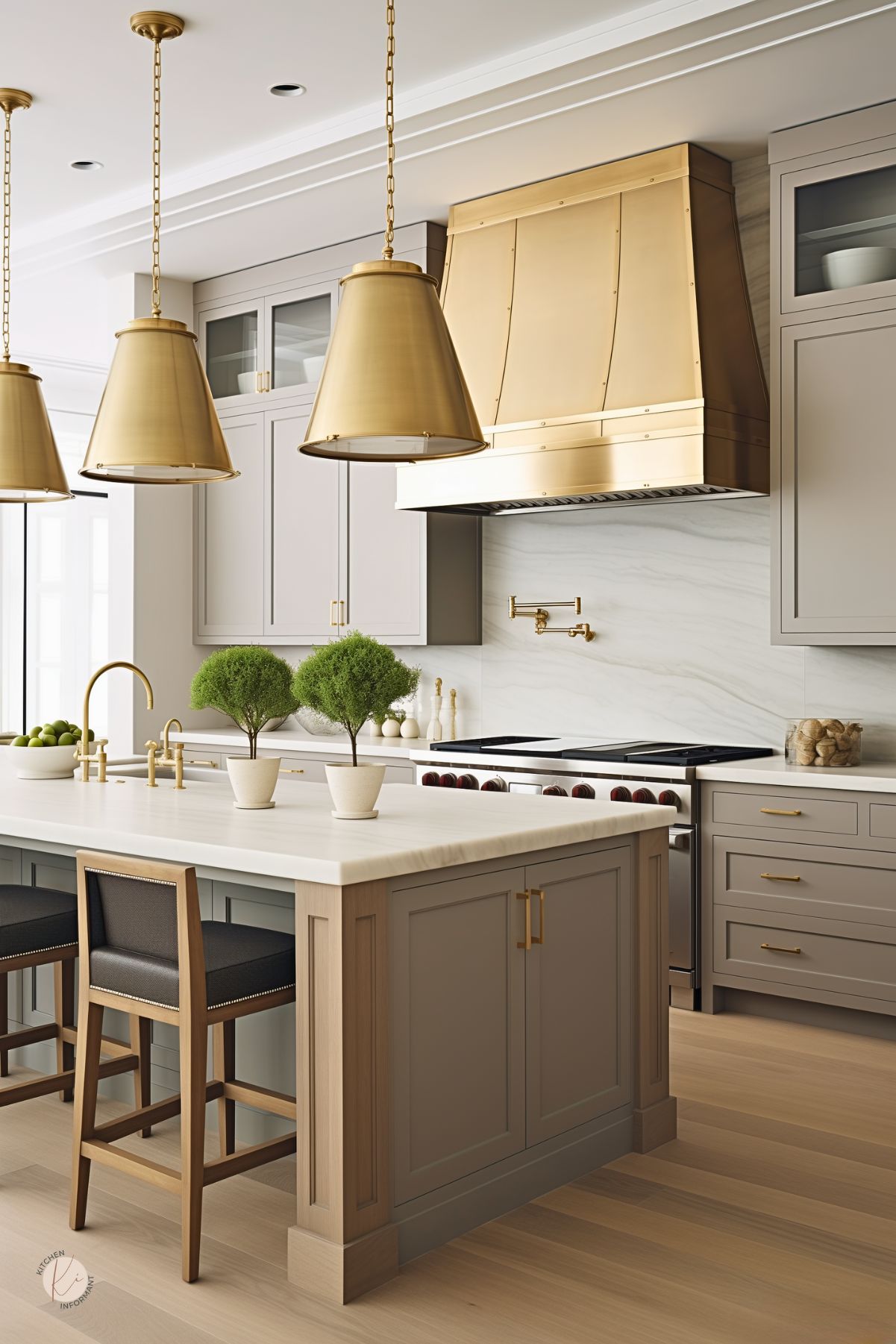 A taupe kitchen with bold brass accents, featuring a statement brass range hood and matching pendant lights. The island has a marble countertop, brass hardware, and wood details, complemented by black upholstered barstools. A sleek marble backsplash and gold pot filler complete the elegant yet modern design.