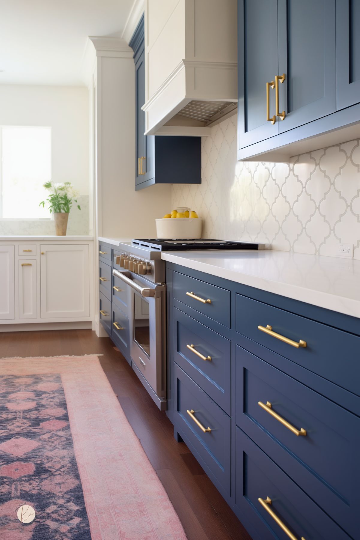 This modern kitchen features navy blue lower cabinets with gold hardware, contrasted by crisp white upper cabinets and a white quartz countertop. A geometric-patterned backsplash adds subtle texture, while a soft pink and blue runner brings warmth to the dark wood flooring. Stainless steel appliances and a sleek range hood complete the polished, contemporary design, with natural light enhancing the fresh, airy feel.