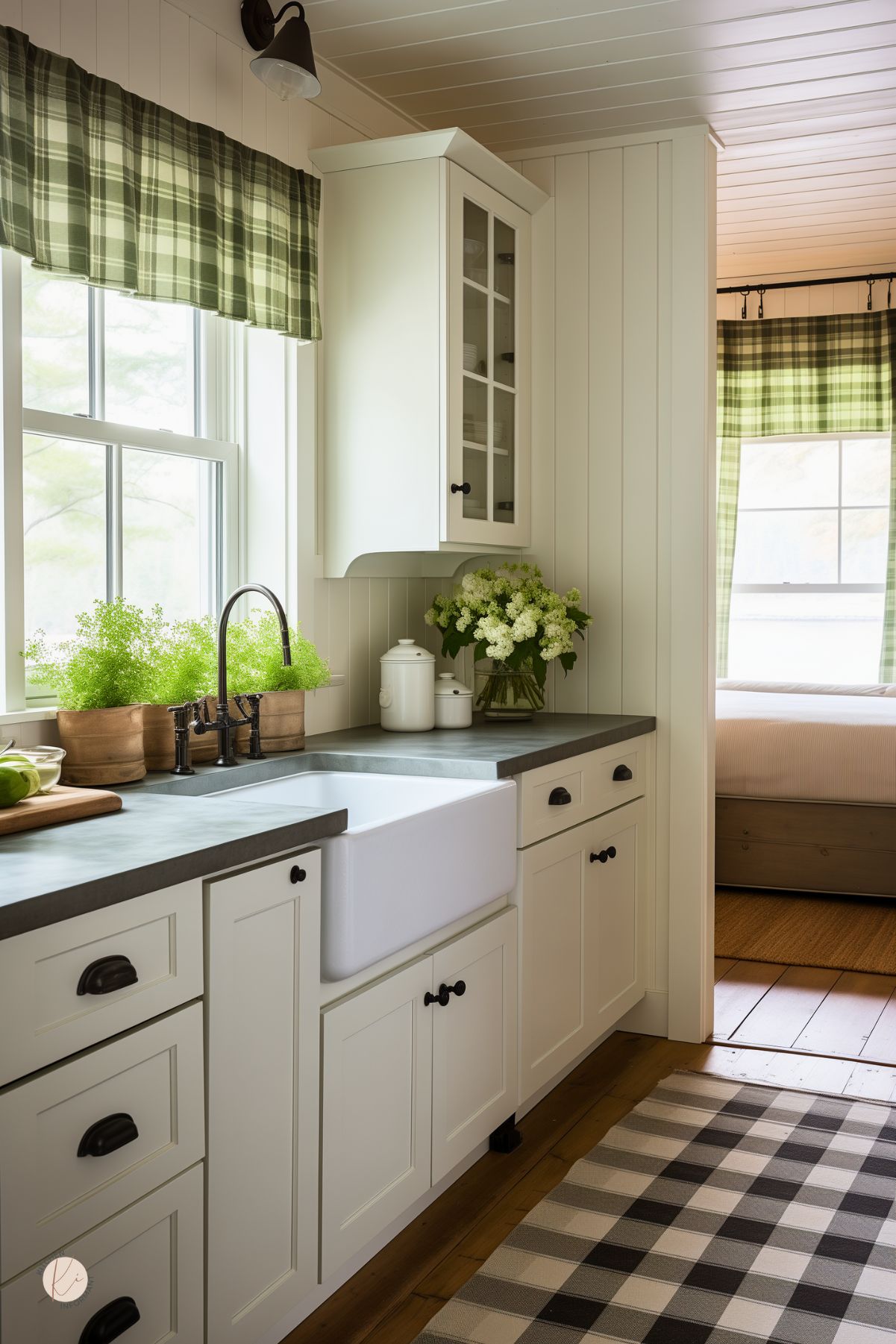 This cozy farmhouse kitchen features cream cabinetry with black hardware, a white farmhouse sink, and dark stone countertops. Green plaid valances and matching curtains in the adjacent room add rustic charm, while a black and white buffalo check rug complements the space. Potted herbs on the windowsill and fresh hydrangeas bring a natural touch, enhancing the warm and inviting ambiance.