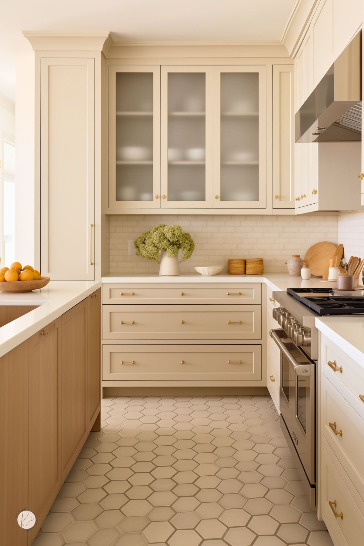 This elegant kitchen features warm cream cabinetry with gold hardware, frosted glass upper cabinets, and a subtle subway tile backsplash. The hexagonal tile flooring adds texture, while the natural wood island provides warmth and contrast. Stainless steel appliances and minimalist decor, including a vase of green hydrangeas and wooden accents, enhance the serene, sophisticated atmosphere.