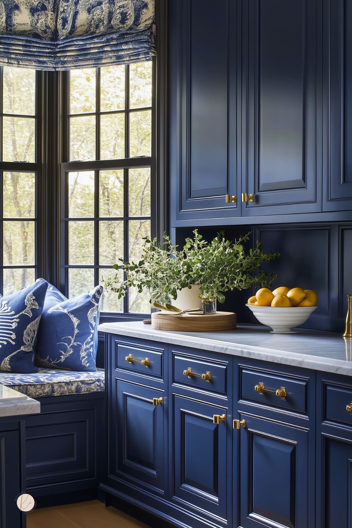 A kitchen with navy blue cabinets, gold hardware, and marble countertops. A built-in window seat with patterned cushions adds charm, while large black-framed windows bring in natural light. Elegant molding and fresh greenery enhance the classic design.