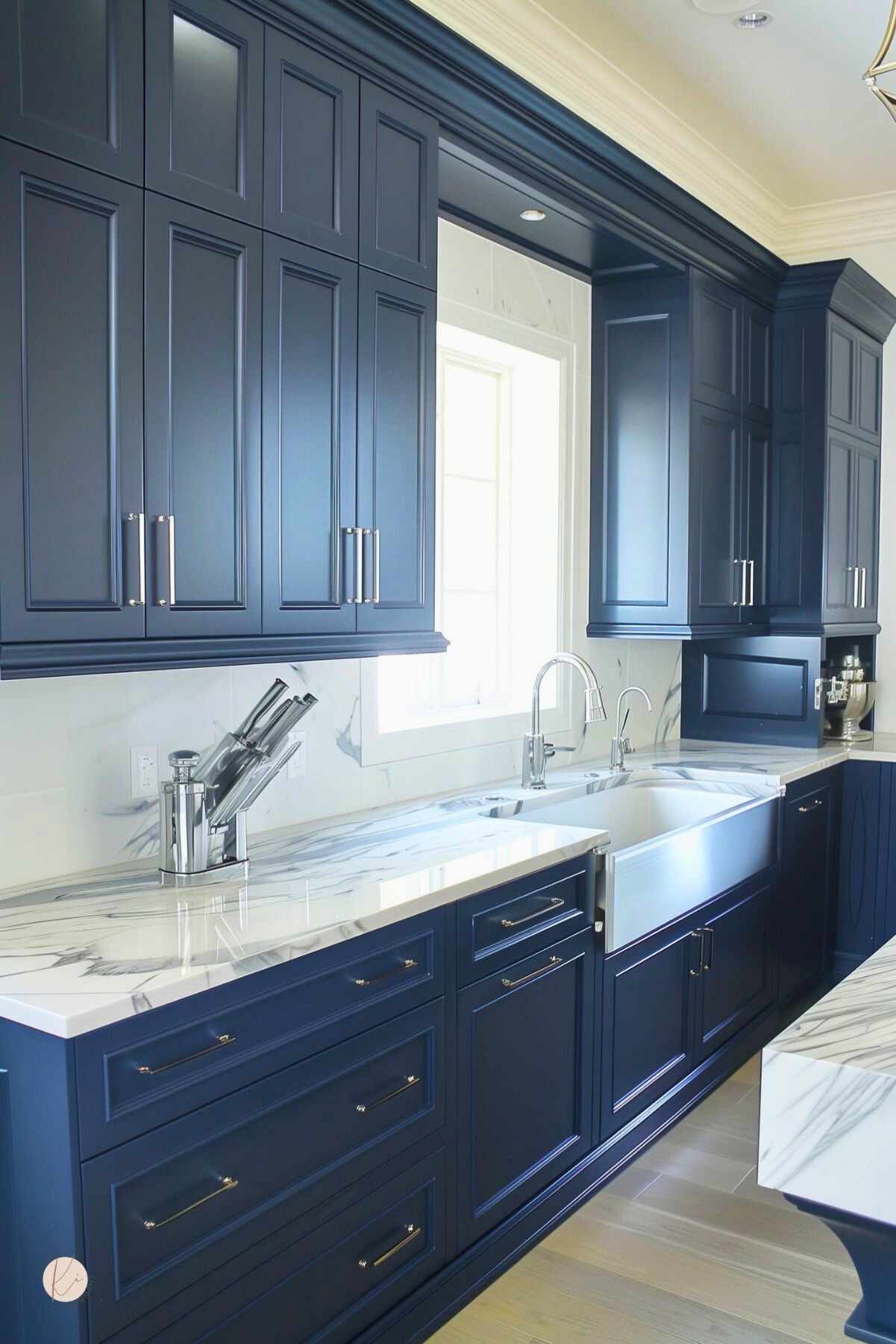 A kitchen with navy blue cabinets, marble countertops, and a stainless steel farmhouse sink. Polished hardware and a marble backsplash add elegance, while natural light enhances the crisp, modern-traditional design.