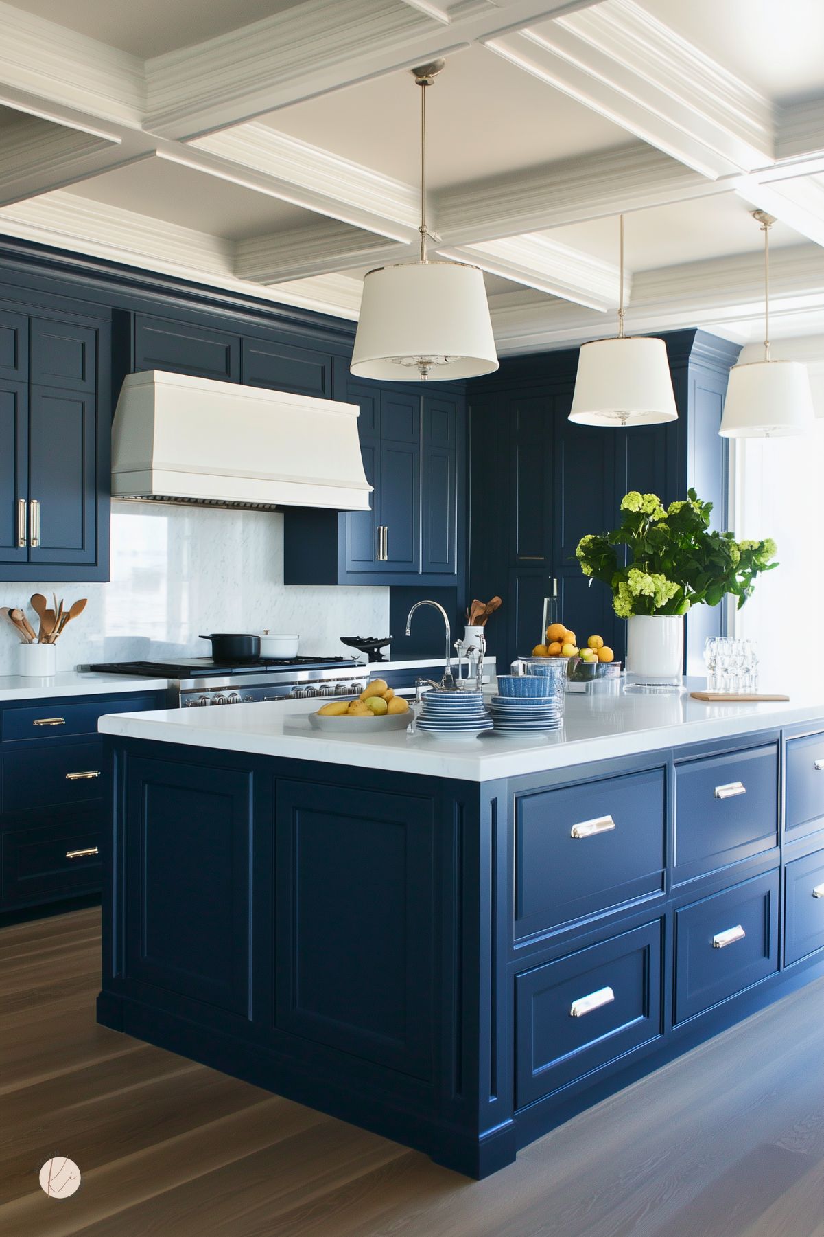 A kitchen with navy blue cabinets, a white quartz island, and a coffered ceiling. A white range hood and marble backsplash contrast with the dark cabinetry, while oversized pendant lights add elegance. Stainless steel hardware and fresh greenery complete the bright, classic design.