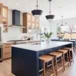 A modern kitchen with a navy blue island, natural wood cabinetry, and a bright open layout. The island features a white waterfall countertop with wooden barstools, complemented by bold navy and gold pendant lights. A navy range hood ties into the design, while glass-front cabinets and a white subway tile backsplash add warmth and contrast. Large windows bring in natural light and outdoor views.