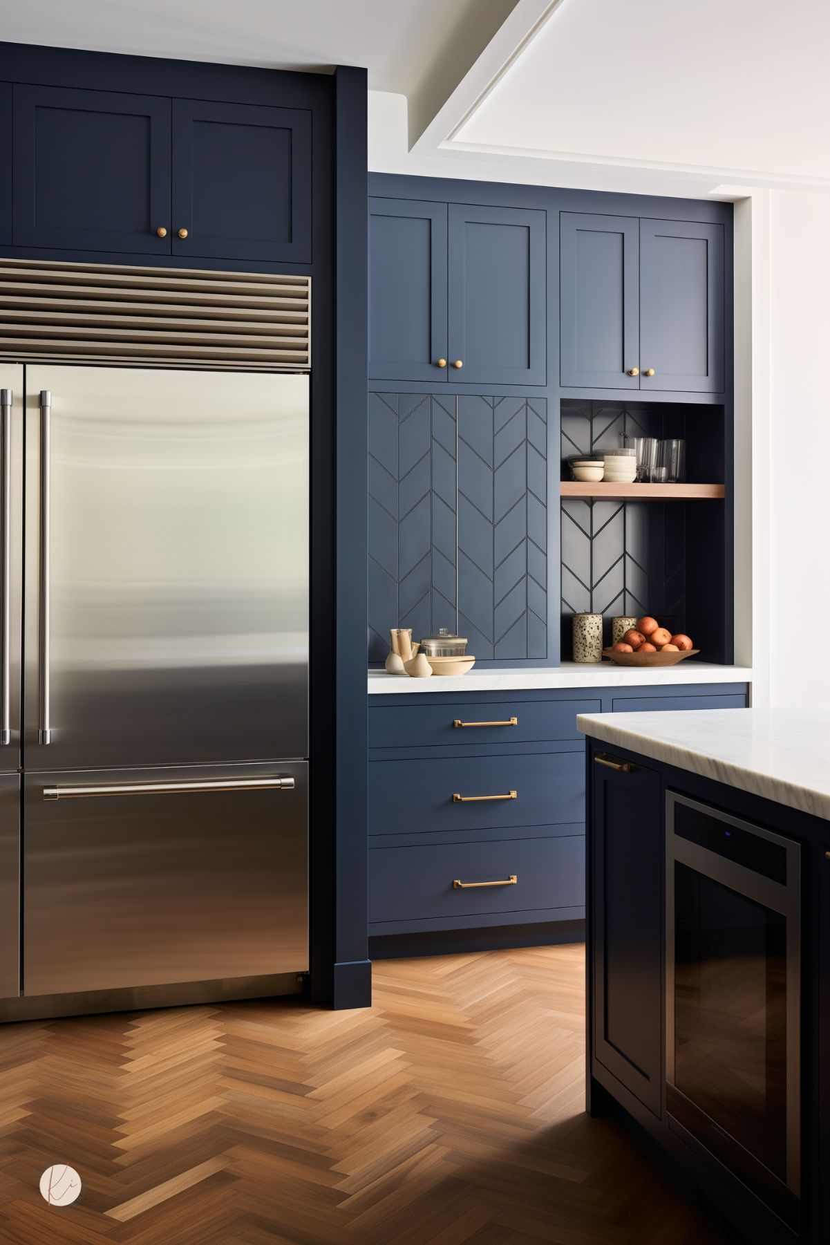 A sophisticated kitchen with navy blue cabinetry, brass hardware, and a chevron-patterned accent wall. A built-in nook with open shelving showcases dishware and warm wood tones. A stainless steel refrigerator adds a sleek contrast, while herringbone wood flooring enhances the space's elegant charm. A white marble island with a built-in wine cooler completes the modern yet timeless design.