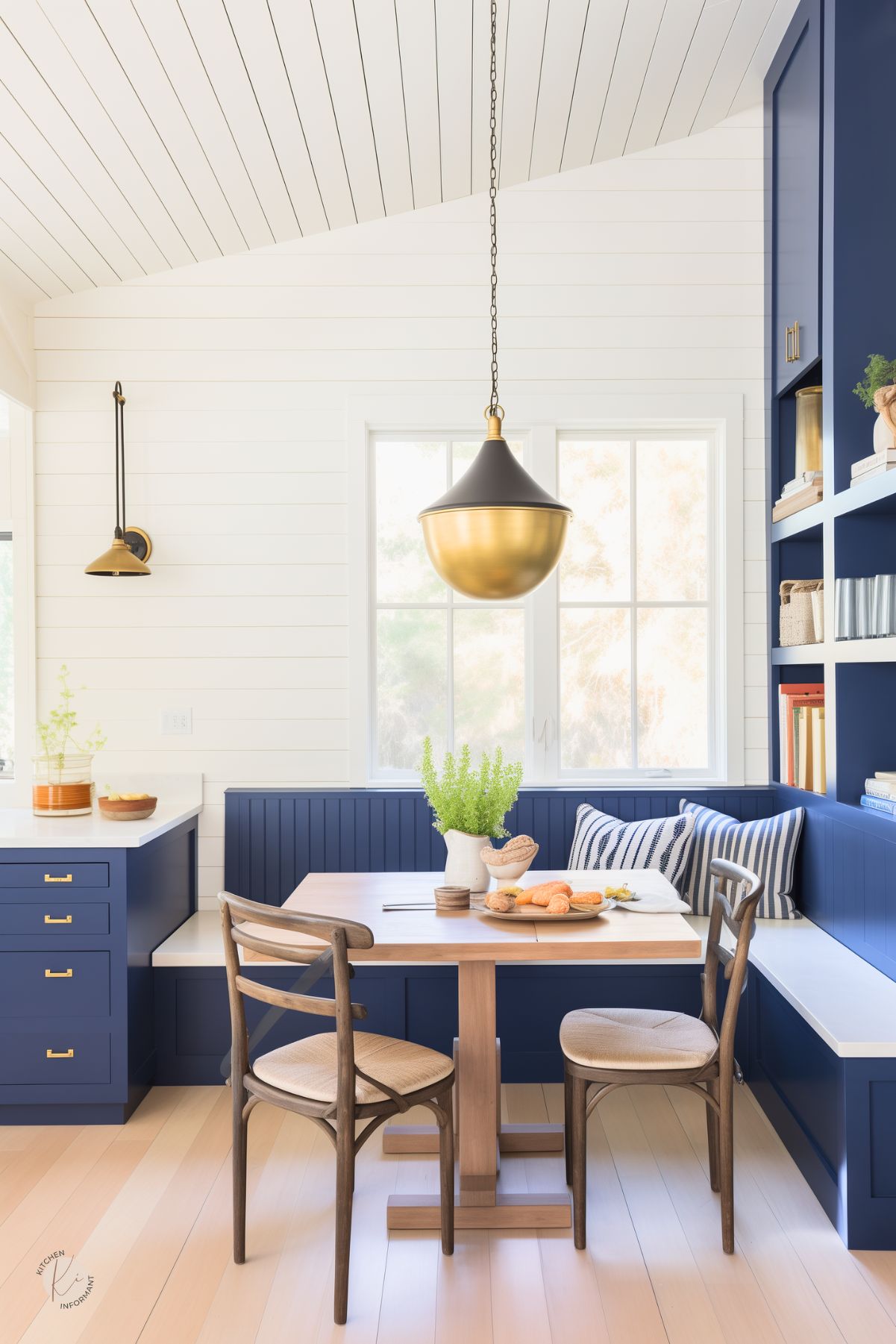 A bright breakfast nook with navy blue built-in seating, natural wood furniture, and a shiplap ceiling. A wooden table is set with pastries and fresh greenery, while striped cushions add a cozy touch. A gold and black pendant light hangs above, complementing the navy cabinetry and brass hardware. Open shelving with books and decor enhances the inviting farmhouse-style space.