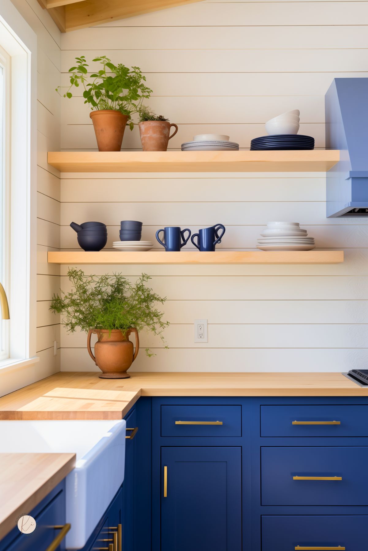 A cozy kitchen with sapphire blue cabinetry, gold hardware, and warm butcher block countertops. Open wood shelving displays blue and white dishware, while potted plants add a natural touch against the white shiplap walls.