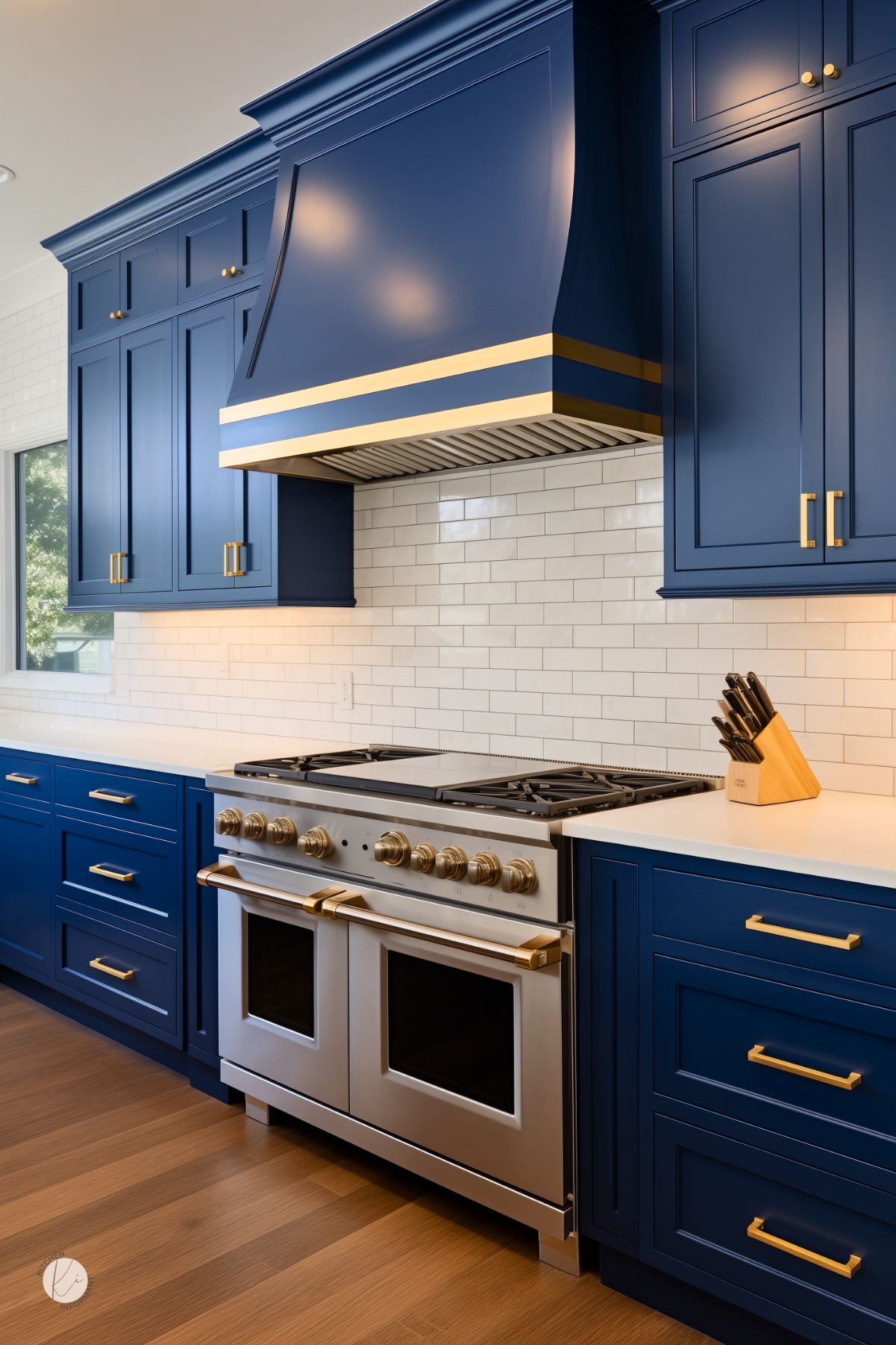 A kitchen with sapphire blue cabinetry, gold hardware, and a matching range hood with gold accents. A stainless steel gas stove sits against a white subway tile backsplash, while white countertops and warm wood flooring complete the design.