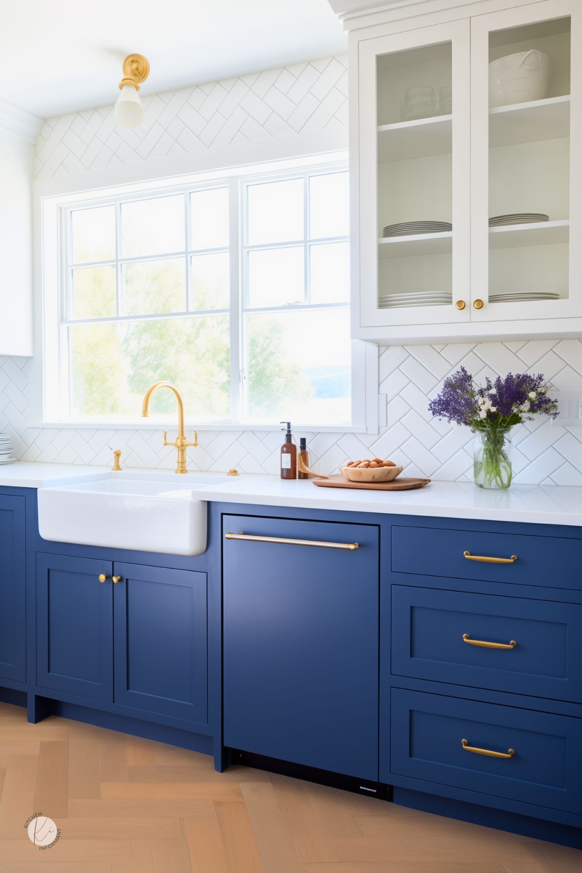 A bright kitchen with sapphire blue lower cabinets, white upper cabinets, and gold hardware. A white farmhouse sink sits beneath a large window, while a herringbone tile backsplash and natural light enhance the airy feel.