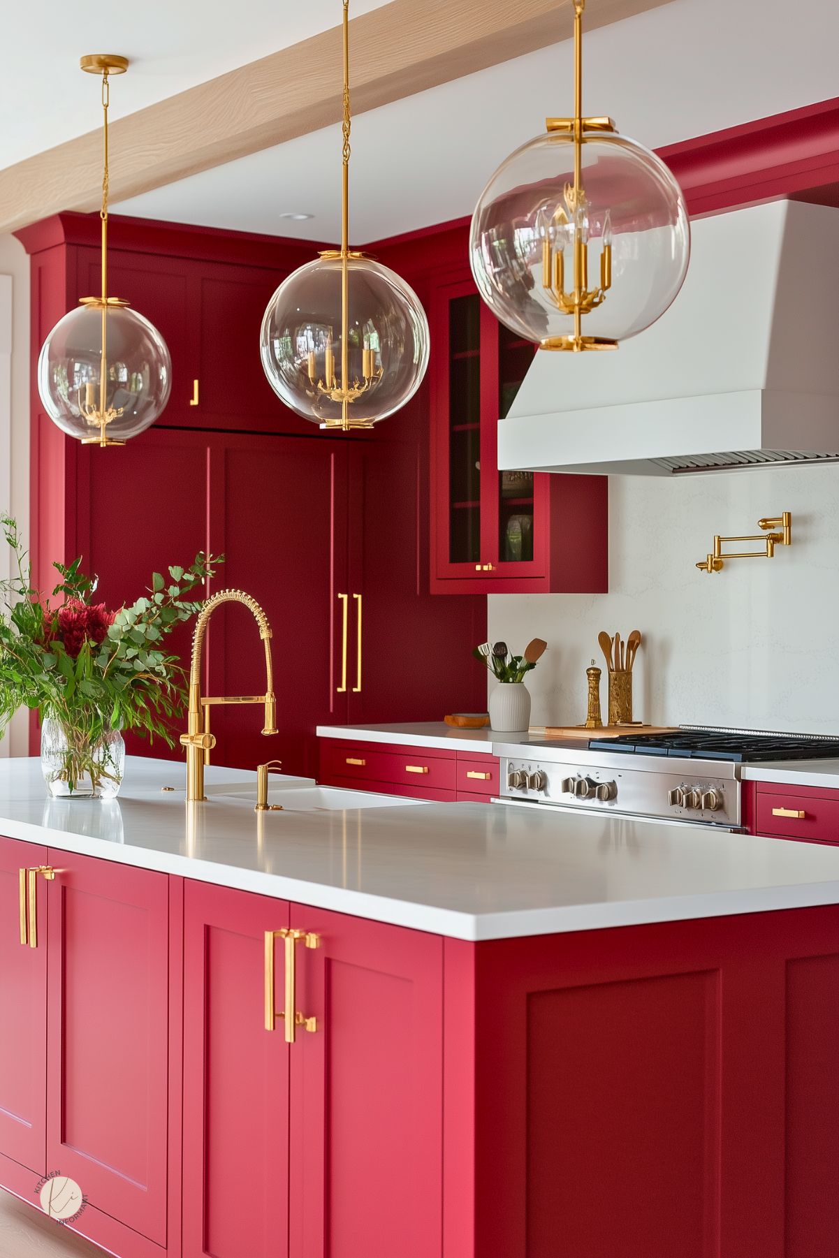 Cherry red kitchen with gold hardware, white countertops, and globe pendant lights. Features a white range hood, brass pot filler, and a large island with a gold pull-down faucet. Decor includes a glass vase of greenery and wood utensils, creating a bold, modern kitchen design.