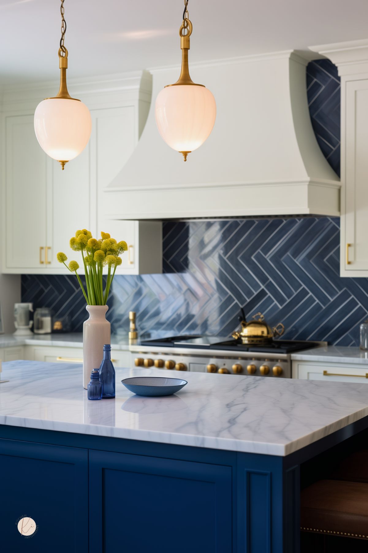 Elegant kitchen with a glossy navy blue herringbone tile backsplash, white cabinetry, and a statement white range hood. A navy blue island with a white marble countertop sits beneath two brass and frosted glass pendant lights. Gold hardware and accents, including a brass teapot and range knobs, add warmth. A vase with yellow flowers and blue glass bottles bring pops of color to the refined, classic design.