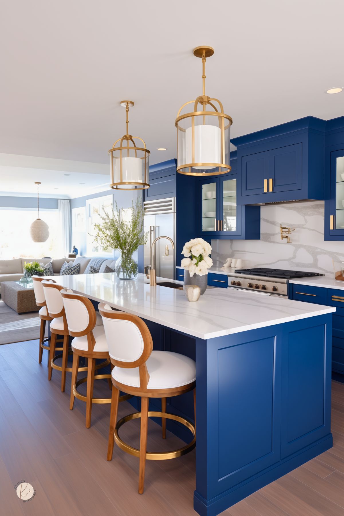 A sophisticated navy blue kitchen with a white marble countertop and gold accents. Elegant pendant lights with gold frames hang above the island, which is lined with white and wood barstools. Glass-front cabinets and a marble backsplash add a luxurious touch, while the open layout flows seamlessly into a bright living space with neutral-toned furnishings.