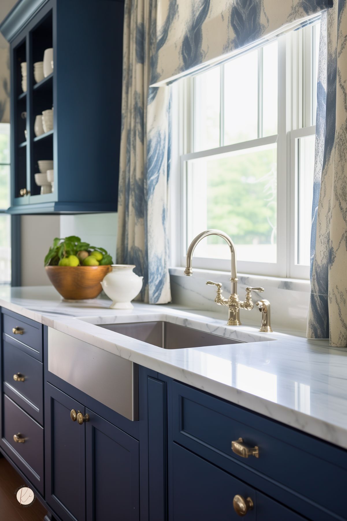 A stylish kitchen with navy blue cabinetry, a white marble countertop, and a farmhouse sink with a polished brass faucet. The window above the sink is dressed with blue and white patterned curtains, allowing natural light to illuminate the space. Open shelving holds neatly arranged white dishware, while a wooden bowl of fresh fruit adds a touch of warmth to the elegant design.