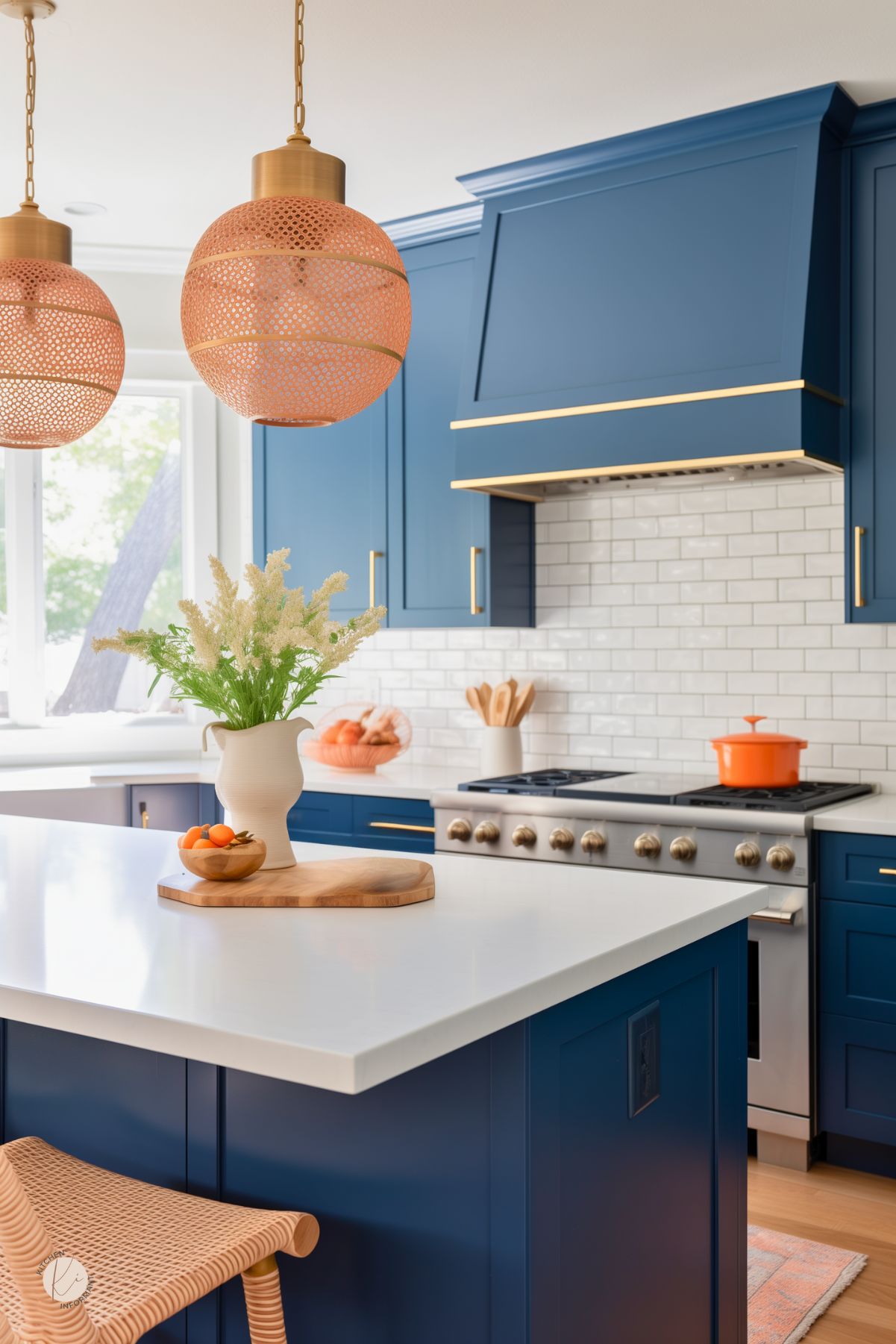 A vibrant kitchen with navy blue cabinetry, white quartz countertops, and brass hardware. The backsplash features classic white subway tiles, while woven rattan pendant lights add warmth. A white vase with fresh greenery sits on the island, along with a wooden cutting board and a small bowl of citrus fruit. The gas range is accented by a bold orange pot, bringing a pop of color to the coastal-inspired space.