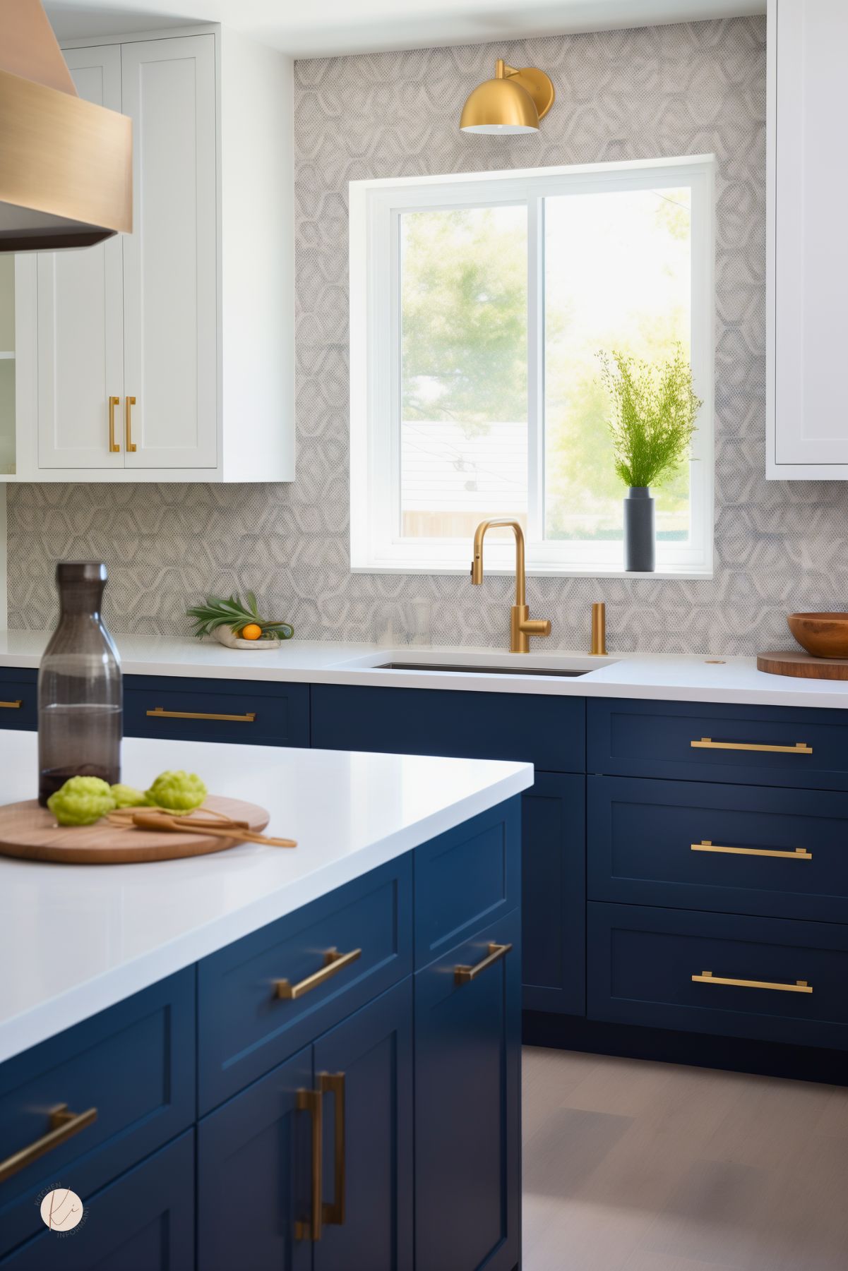 Sophisticated coastal kitchen with navy blue lower cabinets, crisp white countertops, and gold hardware. Light gray patterned backsplash and white upper cabinets add contrast, while a brass faucet and sconce elevate the space with warm metallic accents. Natural light and subtle greenery complete the breezy, modern beach look.
