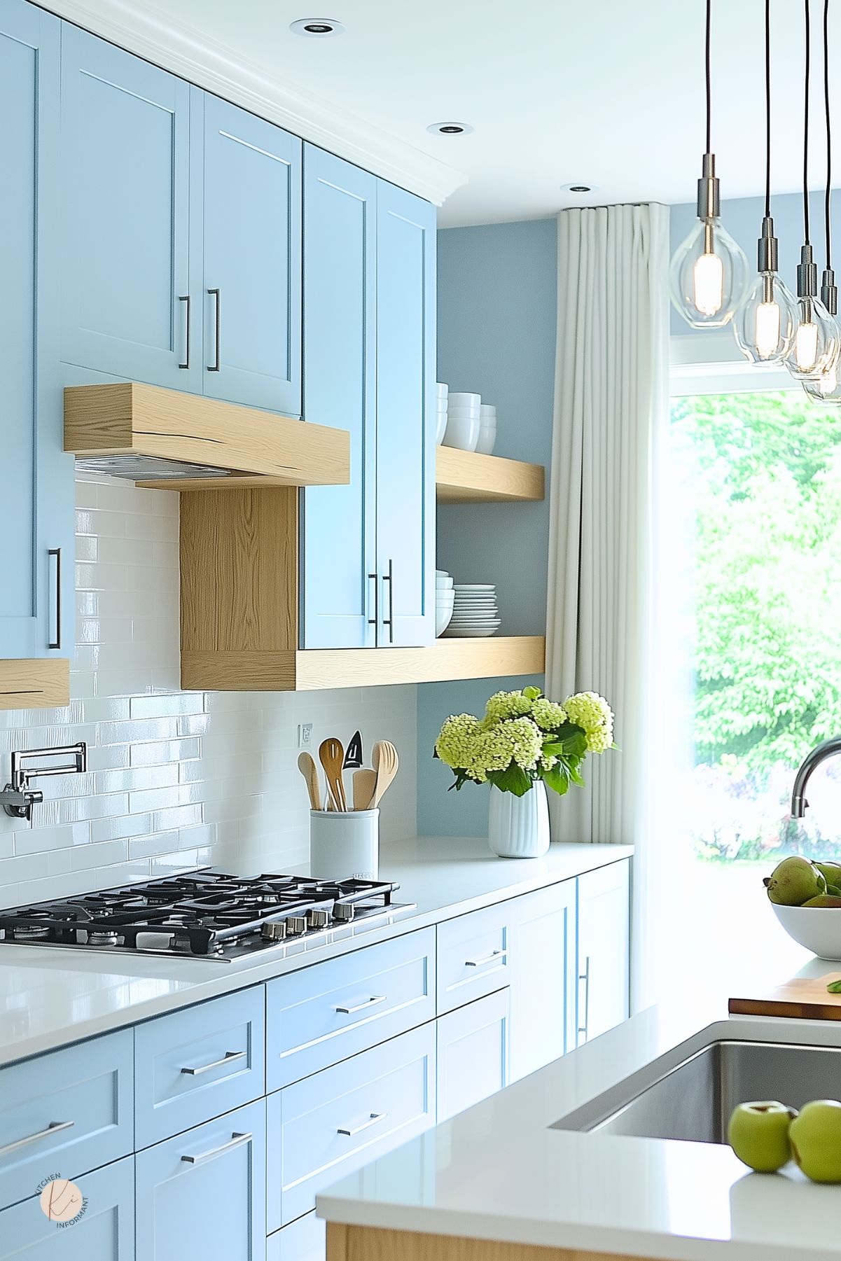 Modern pastel blue kitchen with sleek shaker cabinets, white quartz countertops, and minimalist silver hardware. Light wood open shelving and hood trim add warmth against a glossy white subway tile backsplash. A gas cooktop, vase of hydrangeas, and bowl of green apples add freshness, while hanging Edison bulb lights bring a touch of industrial charm.