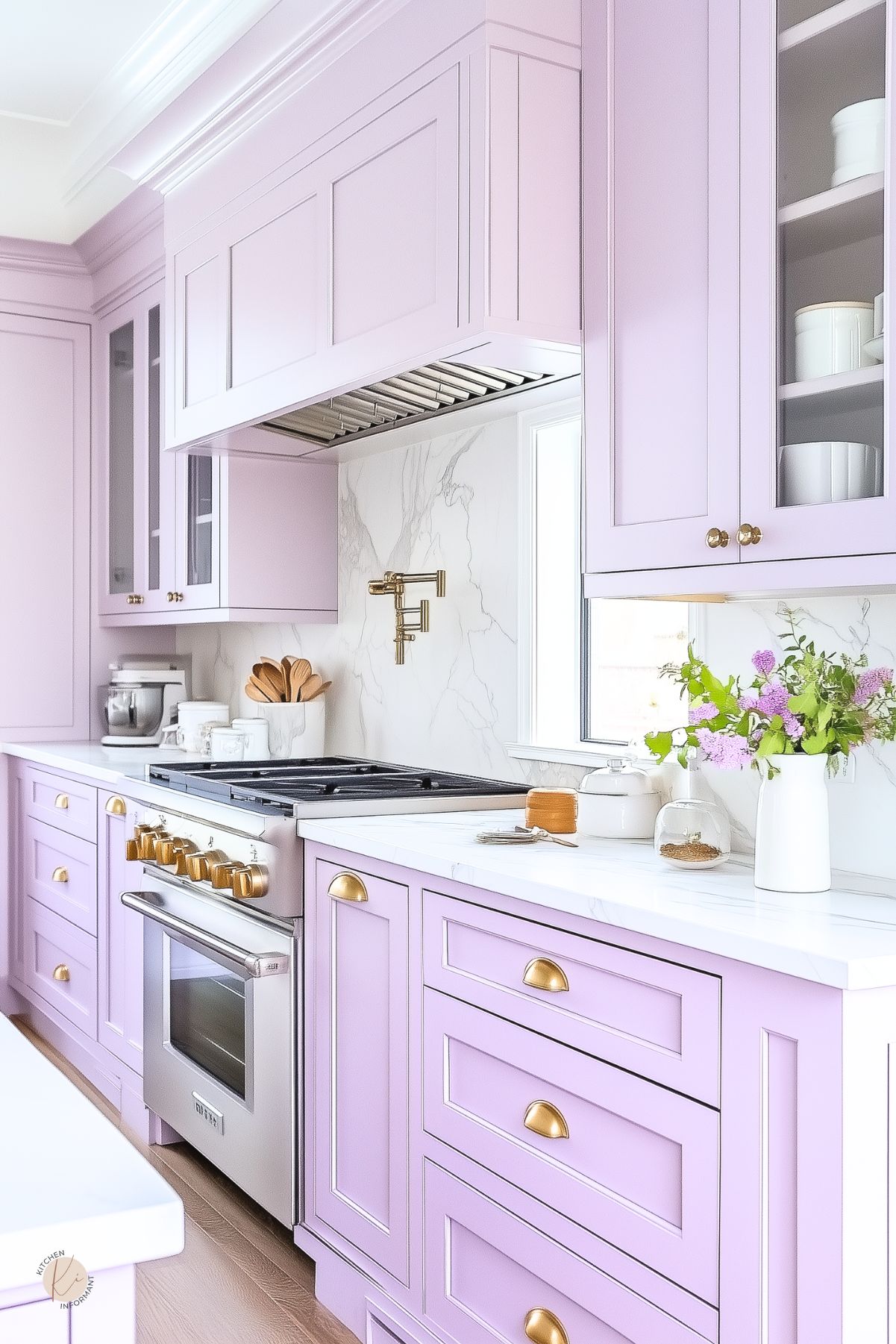 Lavender kitchen with shaker cabinets, white marble countertops, and gold hardware. A sleek stainless steel range with gold knobs sits beneath a marble backsplash and matching pot filler. Glass-front upper cabinets and fresh lilac blooms in a white vase add charm and brightness to the soft, pastel-toned space.