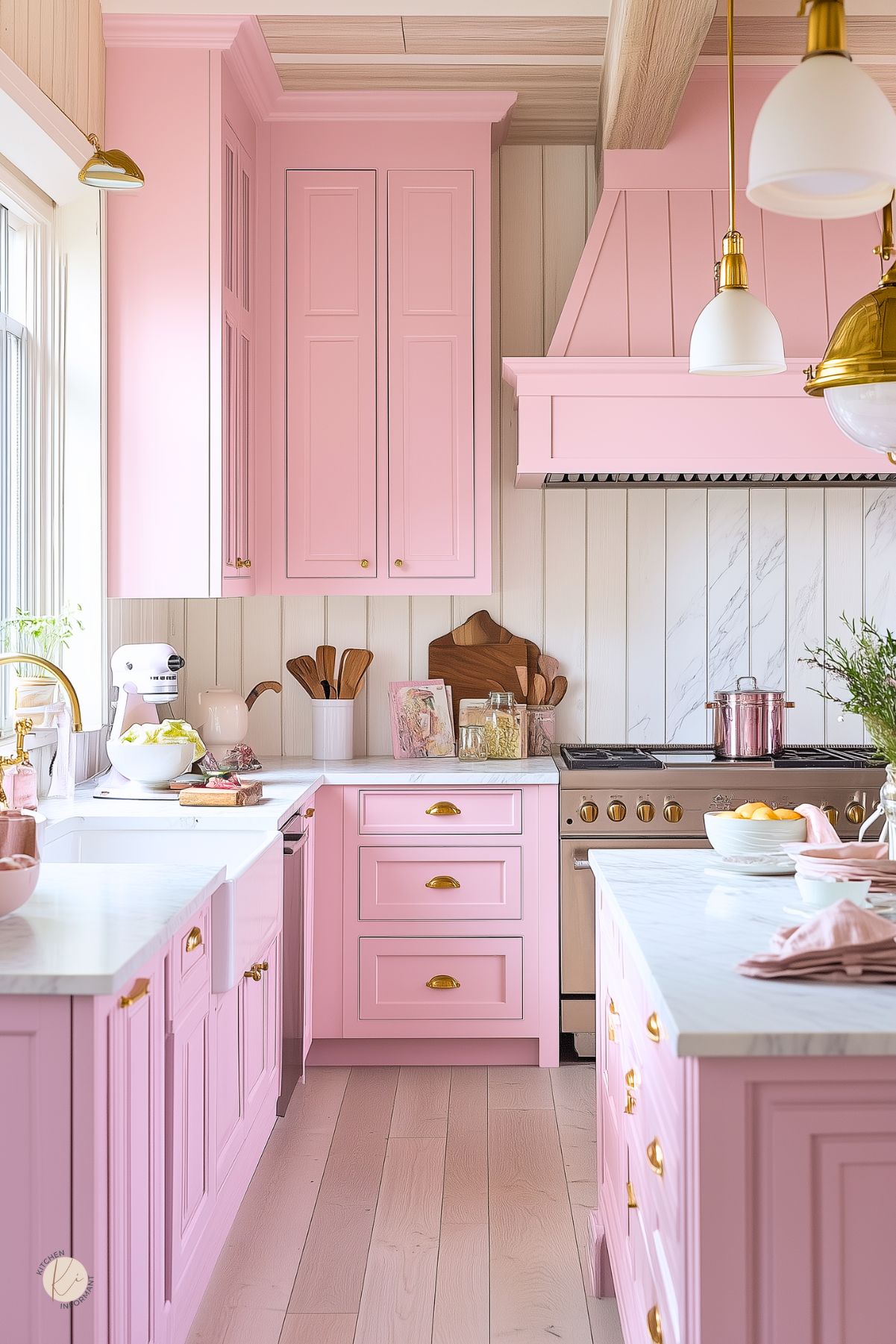 Charming pastel pink kitchen with shaker cabinets, gold cup pulls, and a matching pink range hood. White countertops and backsplash with marble detailing add elegance, while warm wood floors and cutting boards bring balance. Gold-accented pendant lighting and a farmhouse sink complete the playful yet polished cottage-style space.