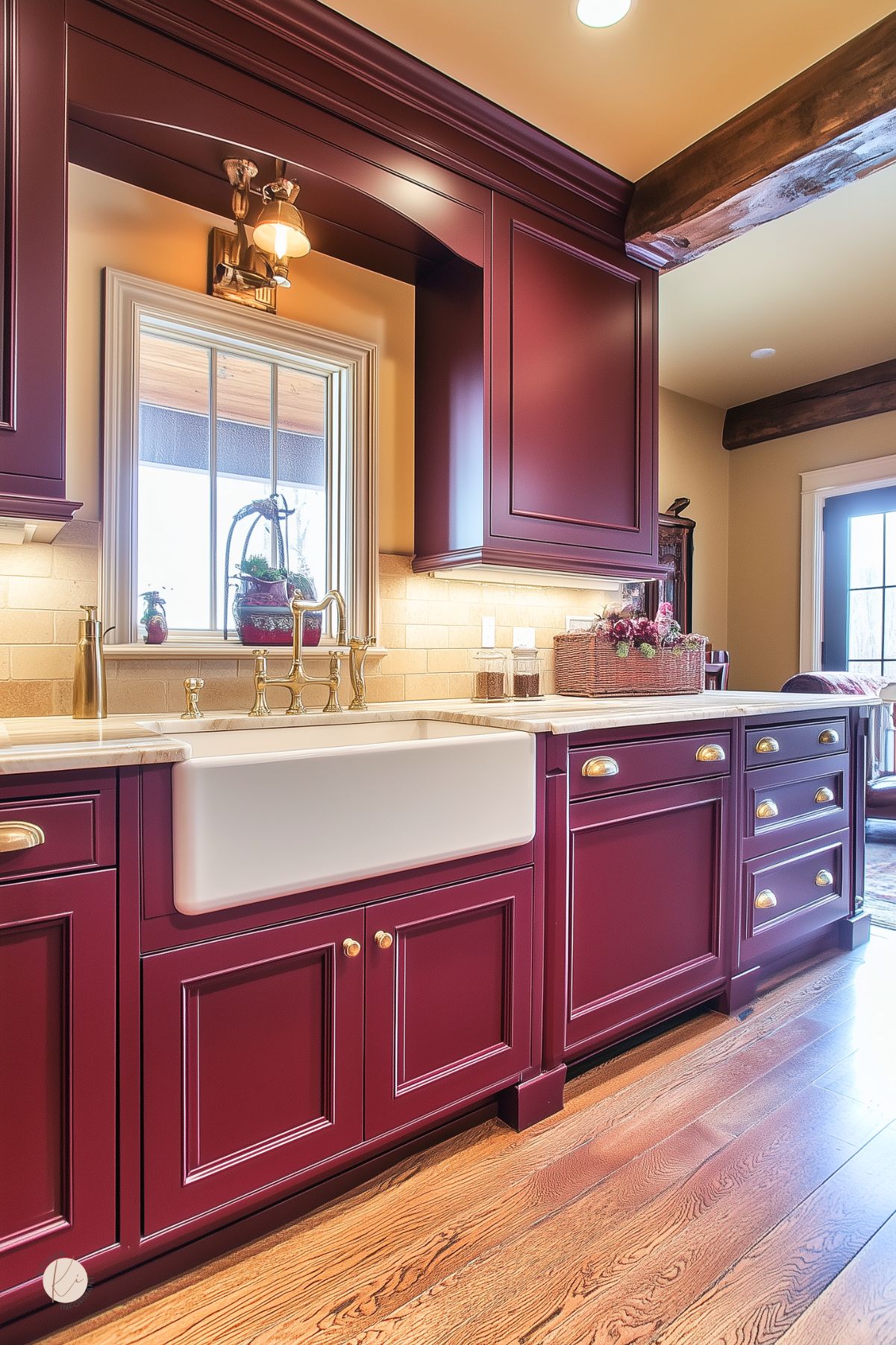 Charming kitchen with deep burgundy cabinetry, a white farmhouse sink, gold hardware and faucet, light stone countertops, rustic wood ceiling beams, and a warm, inviting atmosphere with natural light from a window and glass door.
