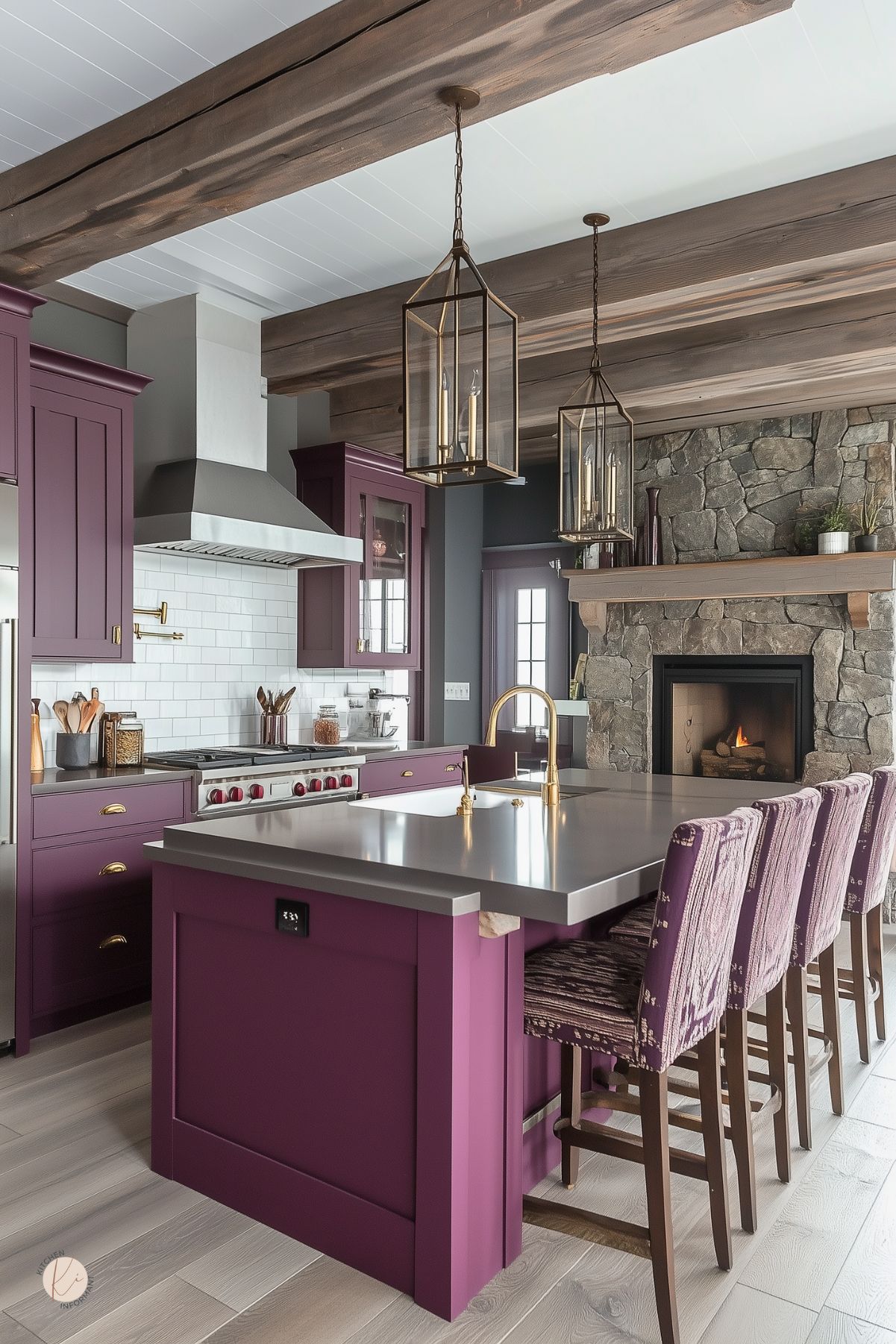 Moody cottage kitchen with rich plum cabinets, brass hardware, and a sleek gray countertop island. Exposed wood beams and large lantern-style pendant lights add rustic charm. A stone fireplace with a wood mantel warms the space, while patterned upholstered barstools complete the cozy, sophisticated look. White subway tile backsplash and a pot filler accent the cooking area.
