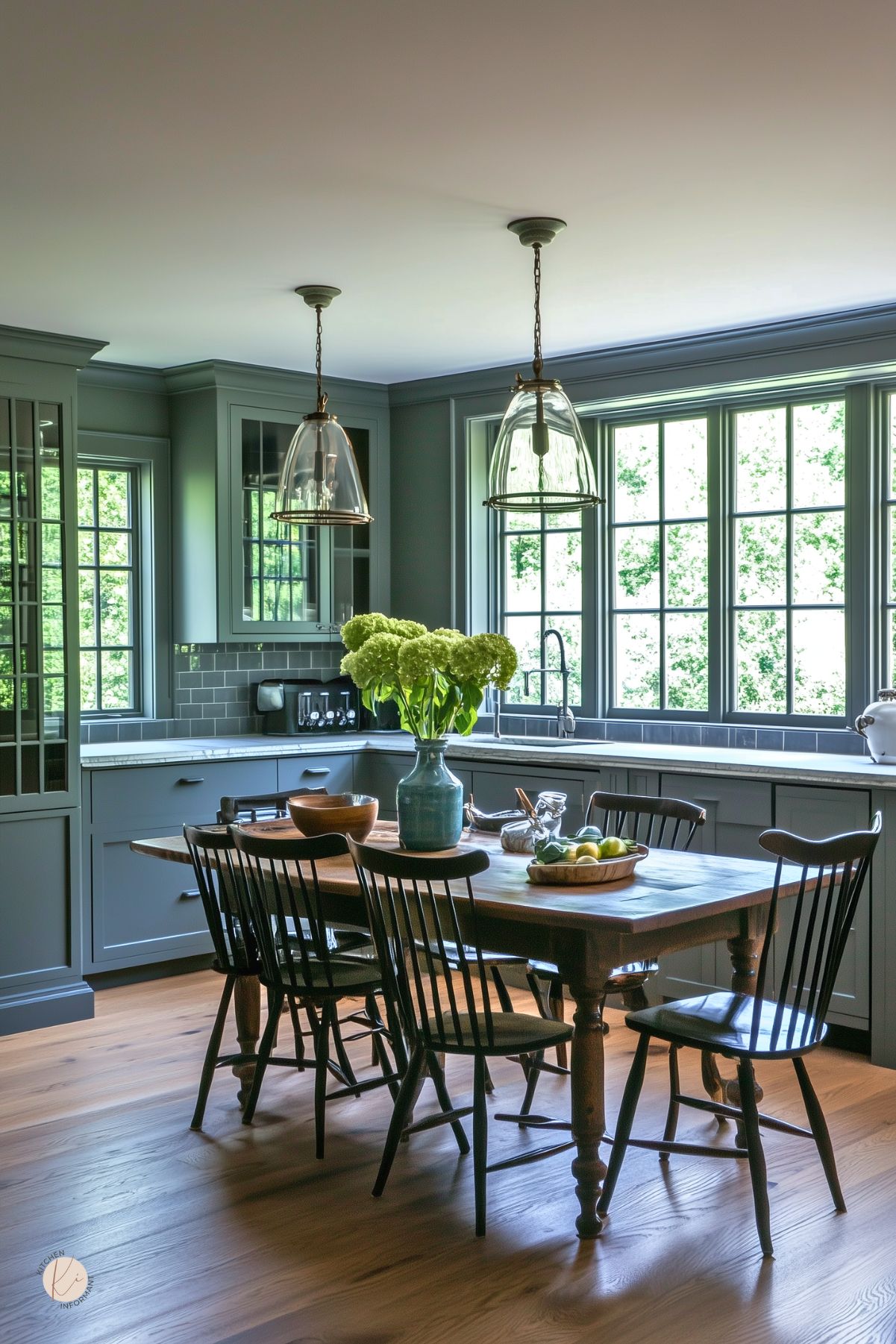 Soft moody cottage kitchen with muted blue-gray cabinetry, subway tile backsplash, and large gridded windows that fill the space with natural light. A wood dining table with black spindle-back chairs anchors the room, topped with a ceramic vase of green hydrangeas. Glass pendant lights and natural wood floors enhance the serene, inviting atmosphere.