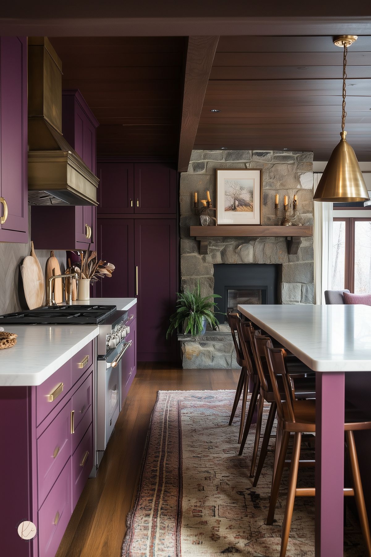 Moody cottage kitchen with deep plum cabinetry, brushed brass hardware, and white countertops. A stone fireplace with a wood mantel anchors the cozy space, complemented by a vintage rug and wood-paneled ceiling. Gold pendant lighting and rich wood accents create a warm, inviting atmosphere with a modern rustic touch.