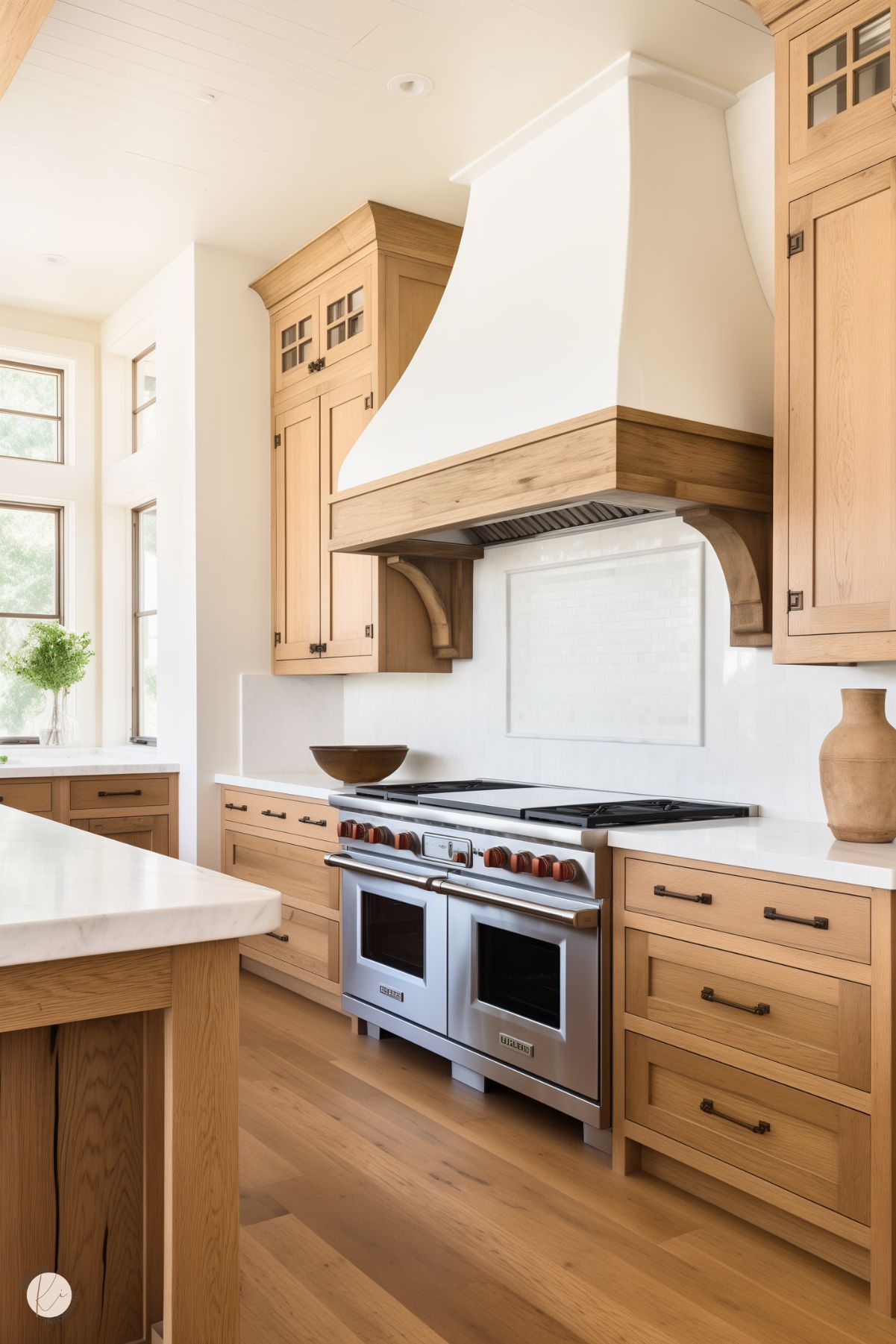 Bright and inviting kitchen with natural white oak cabinets and a statement white plaster range hood accented with oak trim. The space features a commercial-style stainless steel range with wooden knobs, complemented by white quartz countertops and a subtle white tile backsplash. Large windows bring in natural light, and simple decor with earthy tones adds warmth and charm.