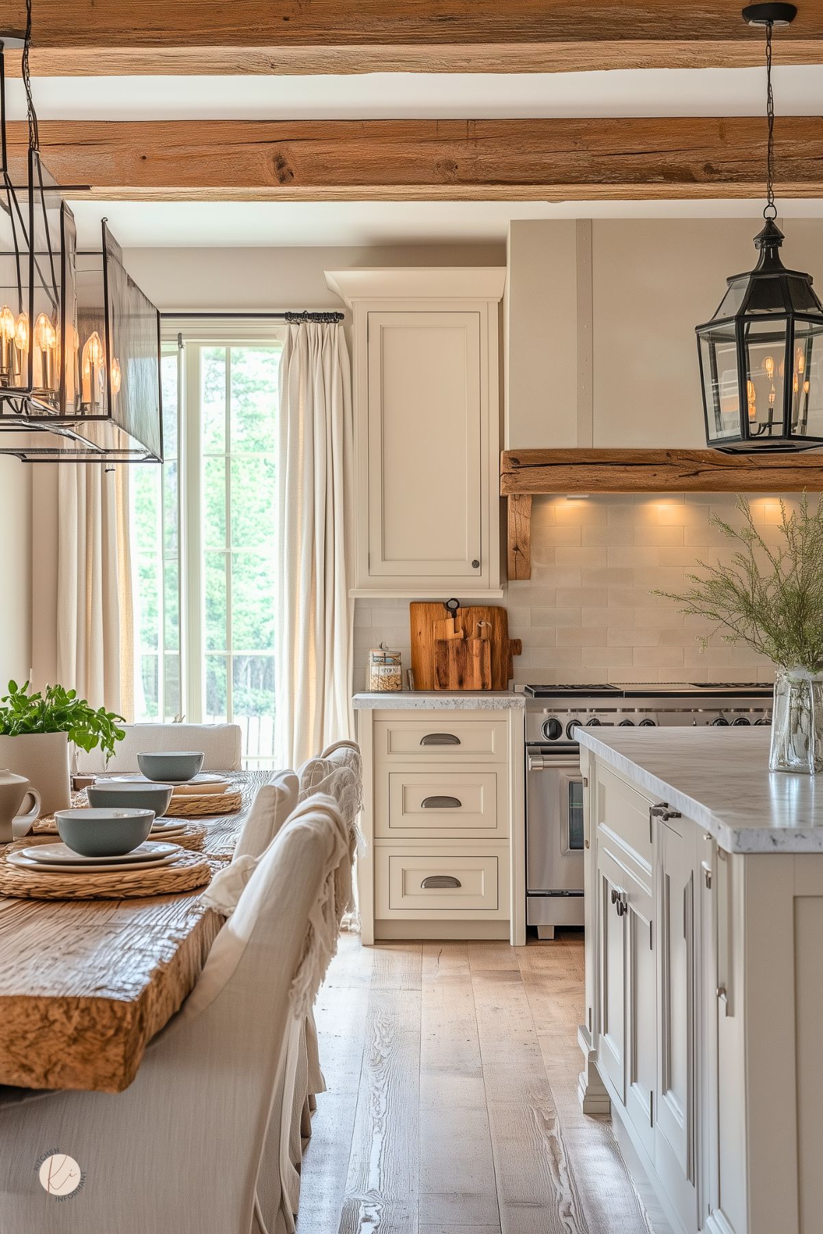 Warm cottage-style kitchen with creamy cabinetry, a rustic wood dining table, and slipcovered chairs. Exposed ceiling beams, woven placemats, and soft linen curtains add to the cozy farmhouse feel. A large lantern-style light fixture and marble countertops bring a touch of modern charm.